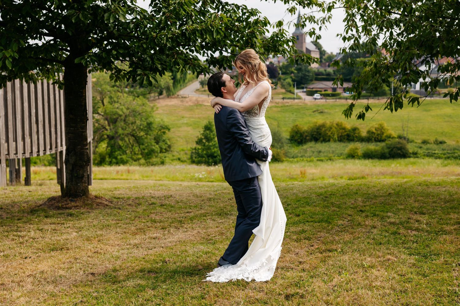 Groom joyfully lifting bride in elegant white gown under a tree in a lush green park.
