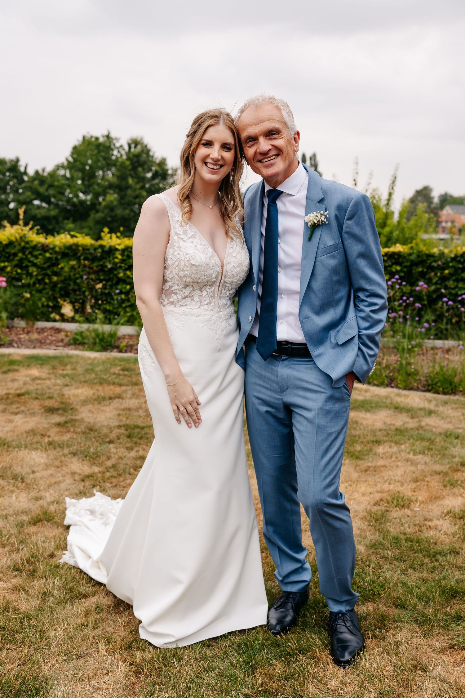 A smiling bride in a white lace gown poses with an older man in a light blue suit at a wedding.