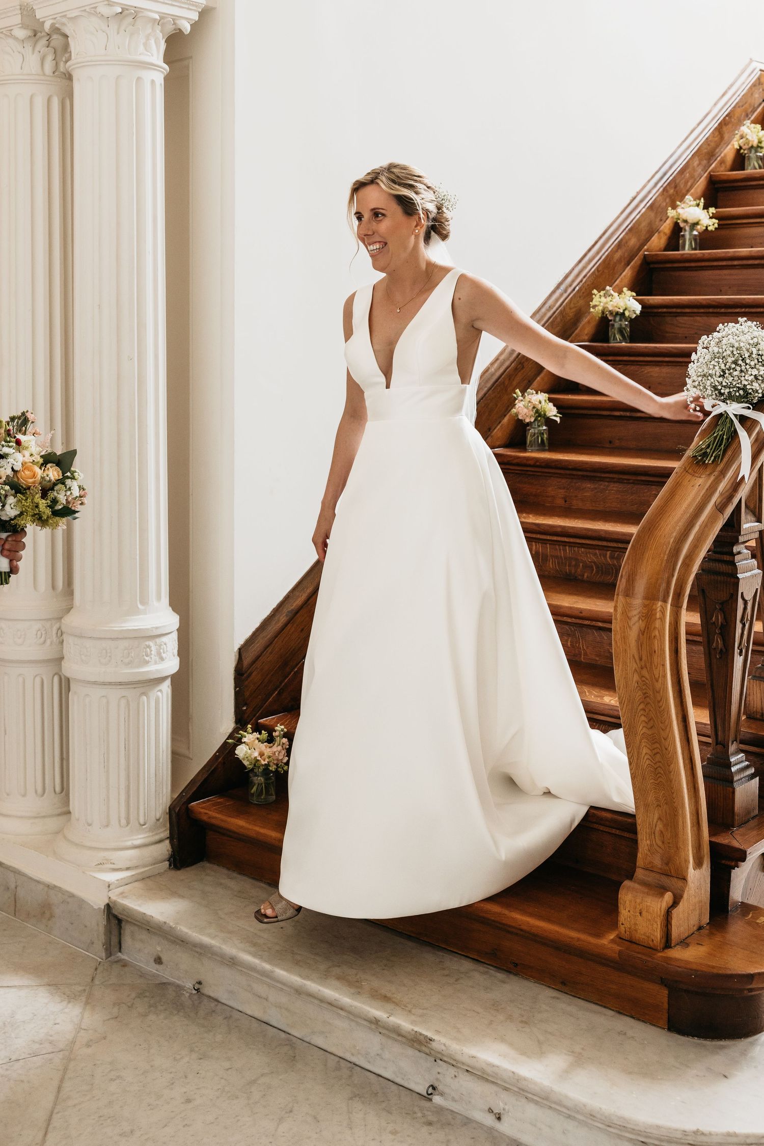 Elegant bride in sleeveless white gown standing on decorated wooden staircase.