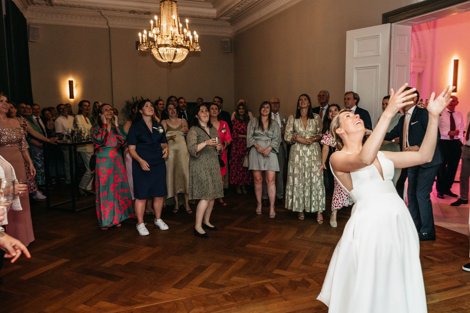 Bride tosses wedding bouquet over shoulder to excited crowd of guests in elegant ballroom.