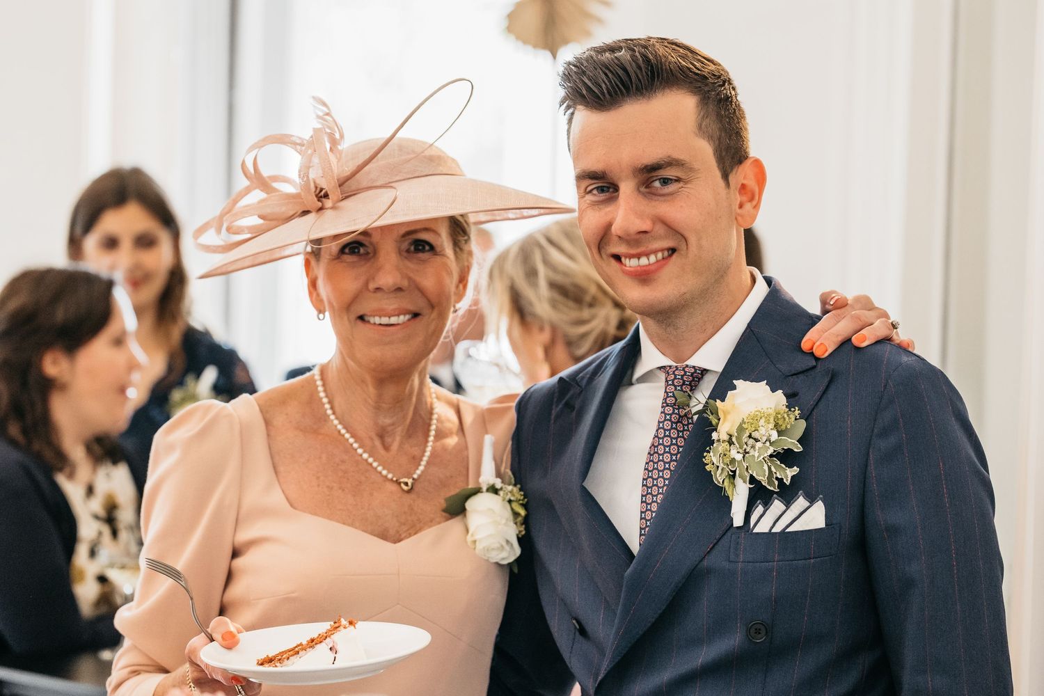 Elegant woman in pink fascinator hat and man in navy suit with boutonniere posing together at a wedding reception.