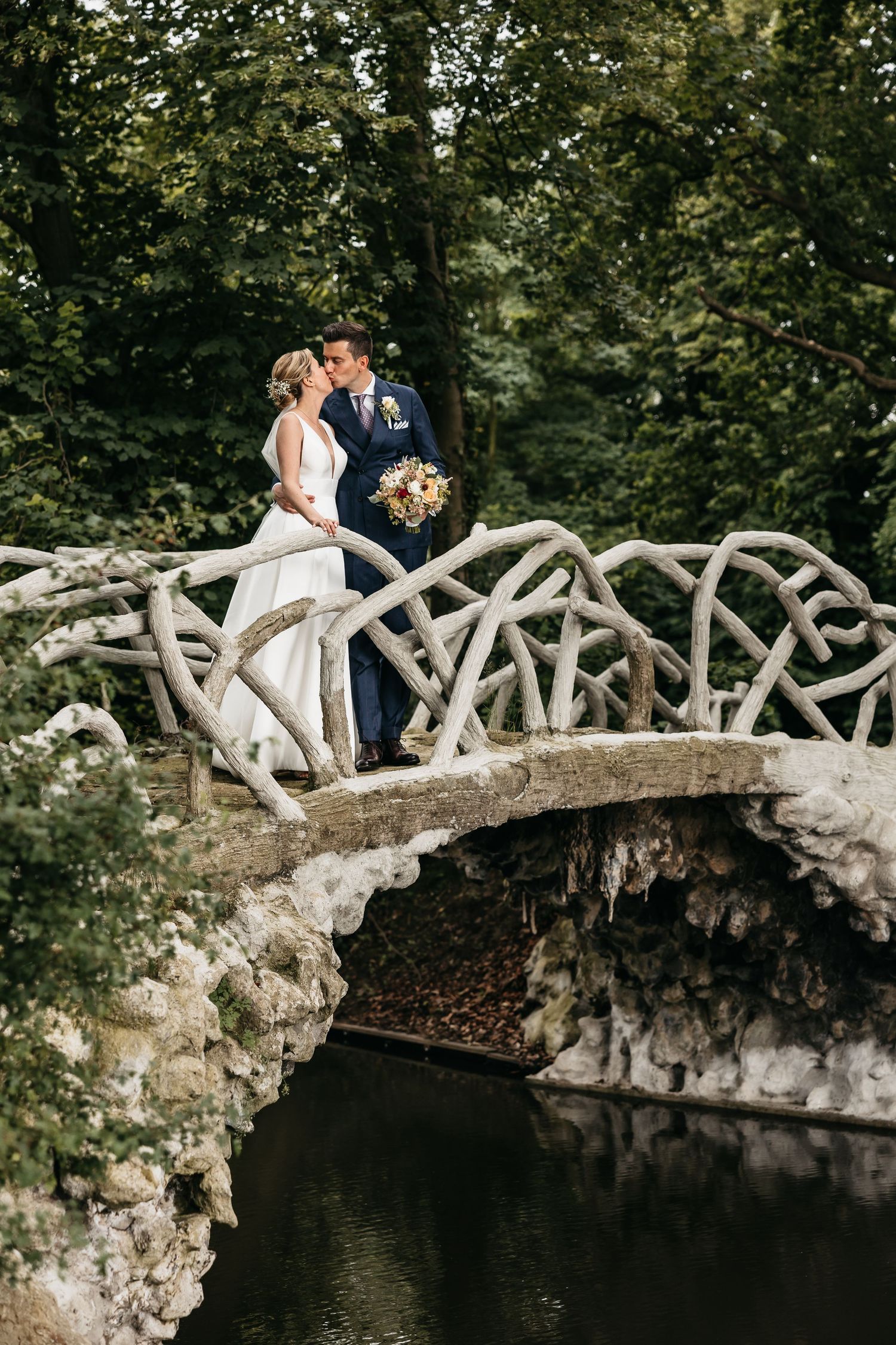 Bride and groom sharing a romantic kiss on an ornate stone bridge in a forest setting.