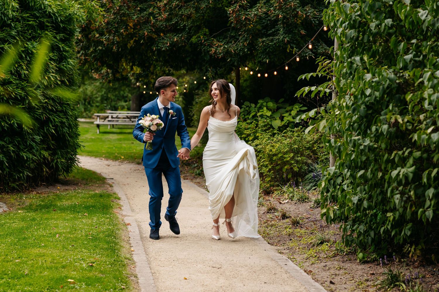 Happy bride and groom running hand in hand along a scenic garden pathway surrounded by lush greenery.