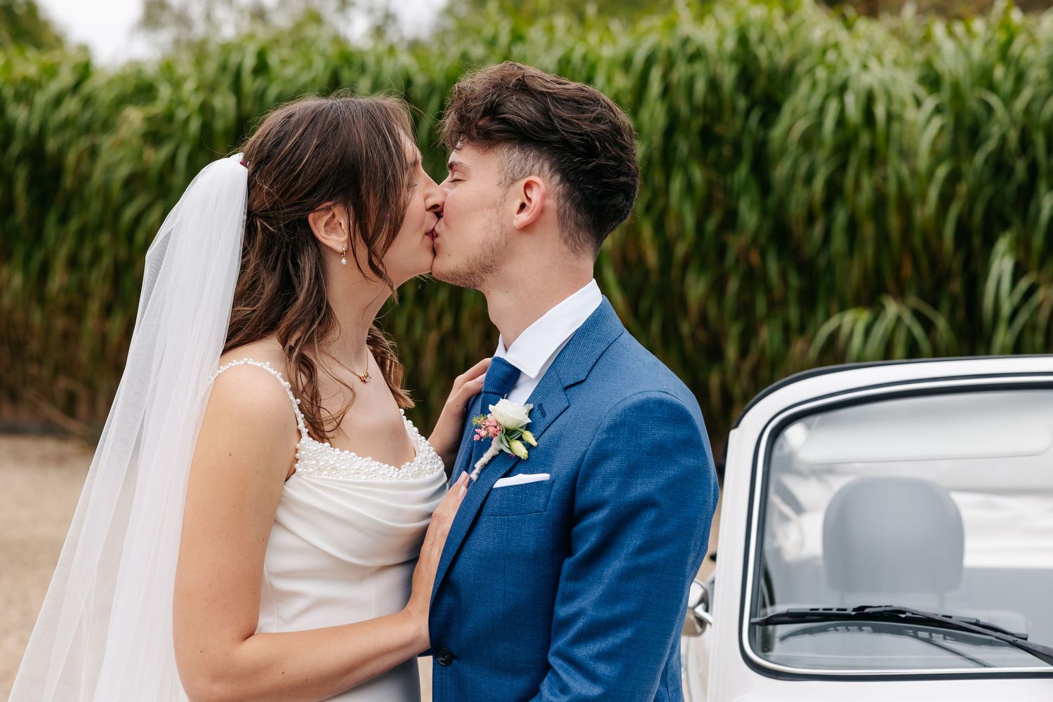 Newlyweds sharing a romantic kiss in front of a vintage white VW Beetle on wedding day.
