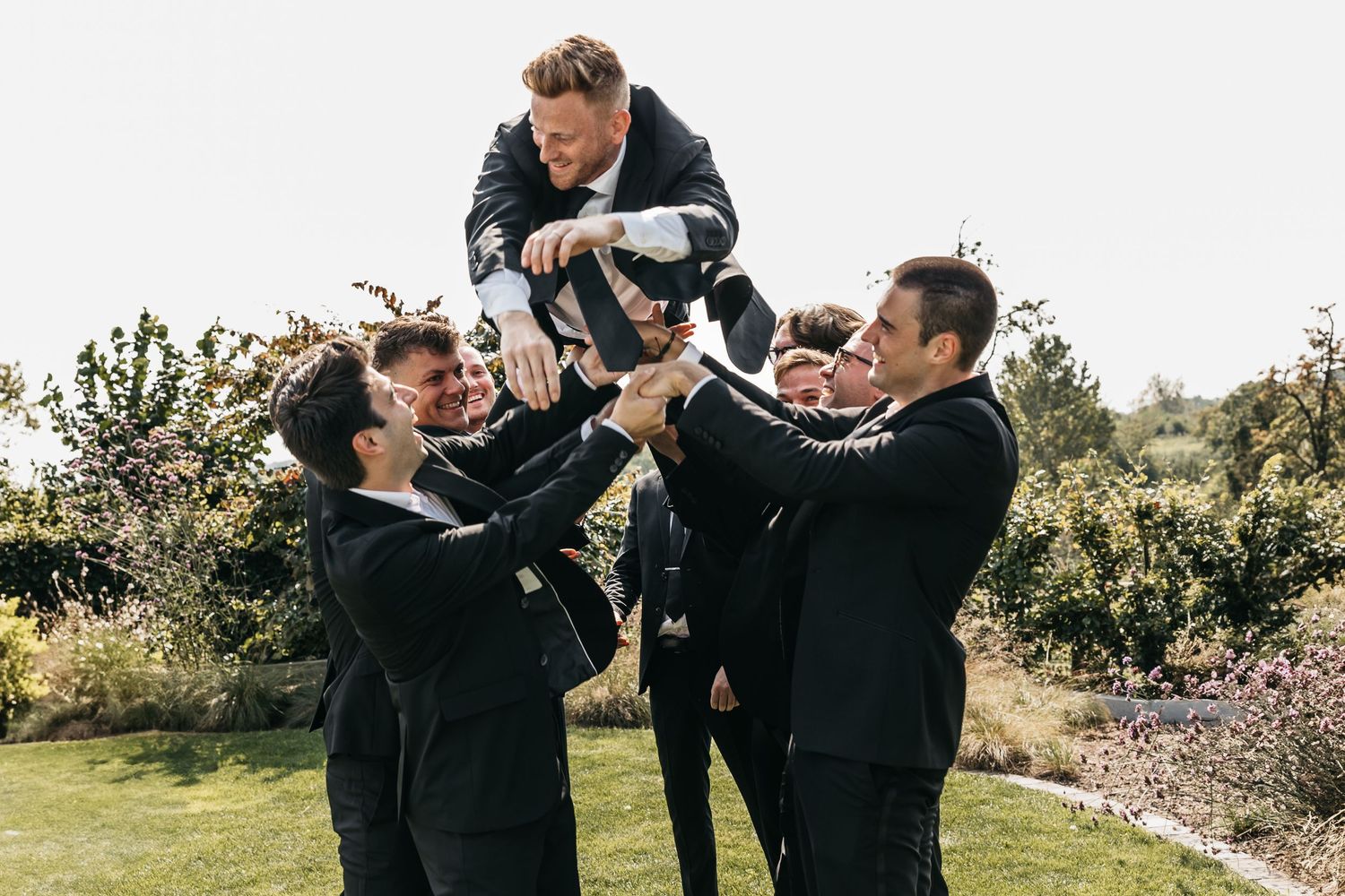 Groomsmen lifting groom into the air during fun outdoor wedding party celebration.