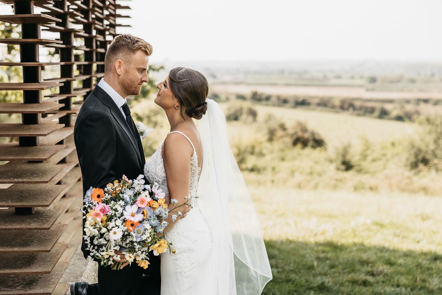 Bride and groom share a romantic moment near a wooden structure with scenic countryside views on their wedding day.
