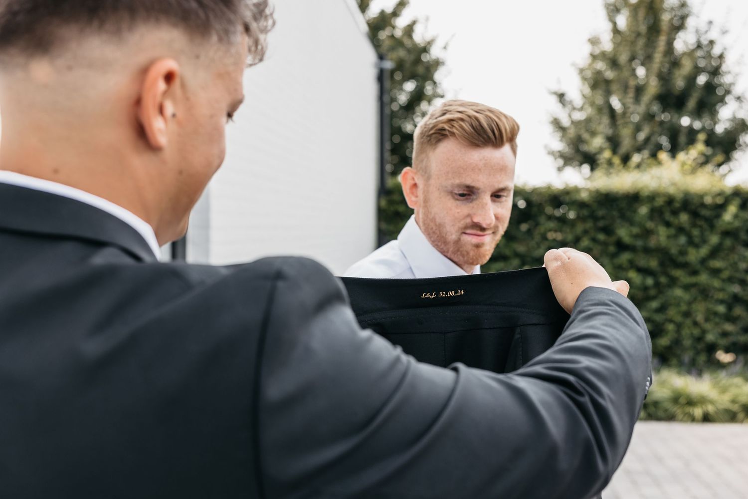 A groomsman helps another man into his suit jacket outdoors before a wedding ceremony.
