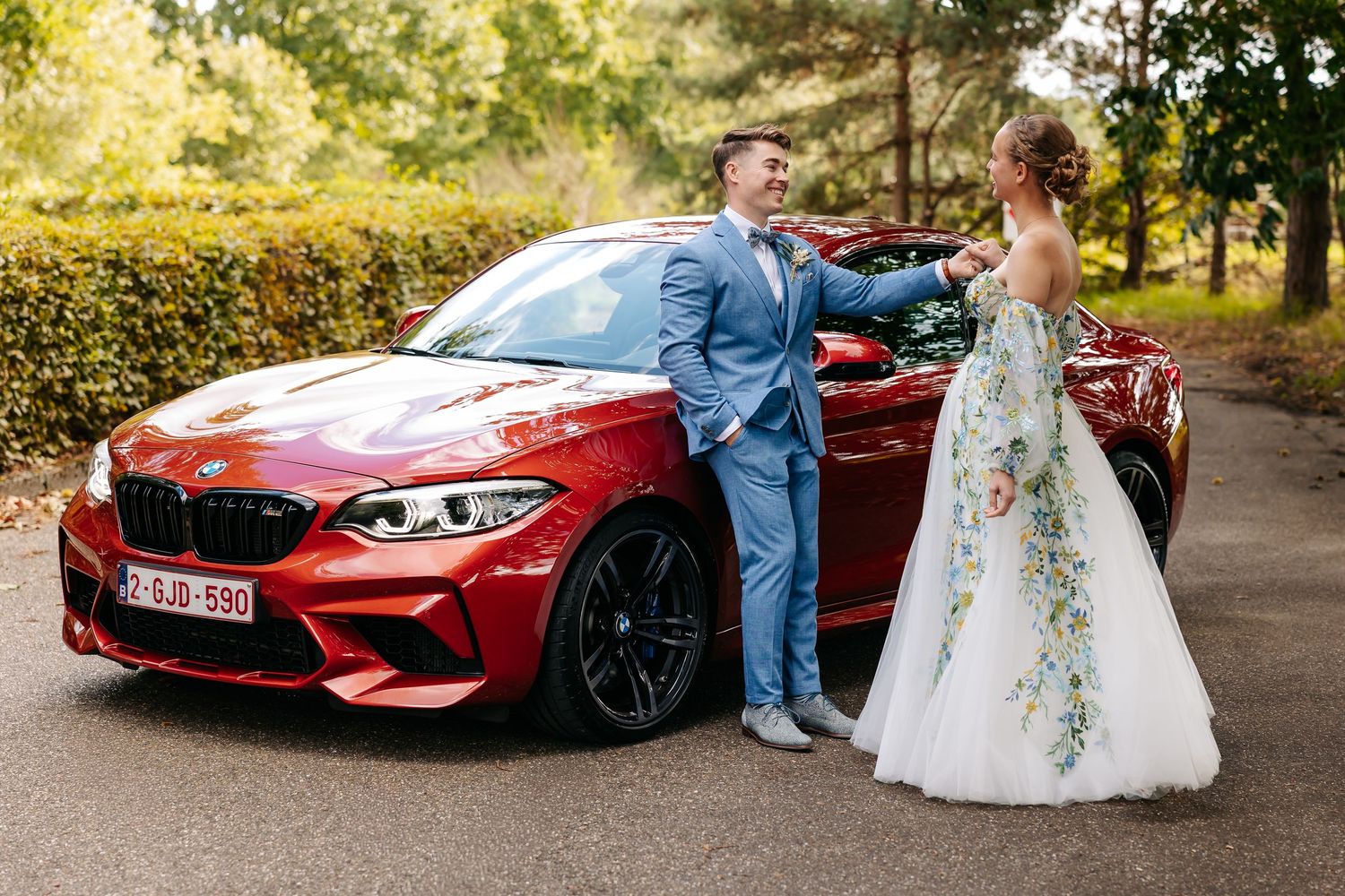 A bride in a floral gown and groom in blue suit pose romantically beside a red BMW M2 wedding car.