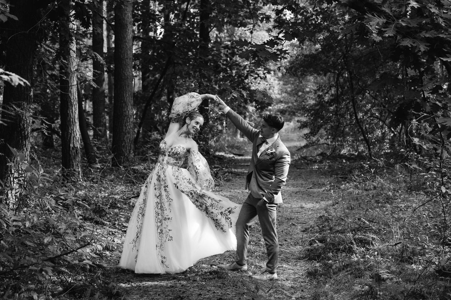 Black and white photo of groom spinning bride in floral dress in forest.