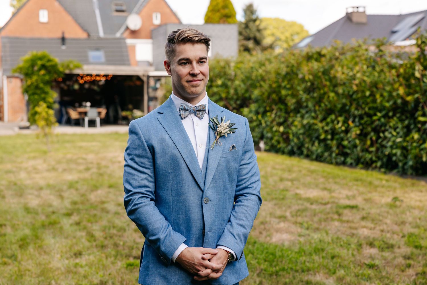 Dapper groom in light blue suit with bow tie and floral boutonniere standing outdoors on lawn.