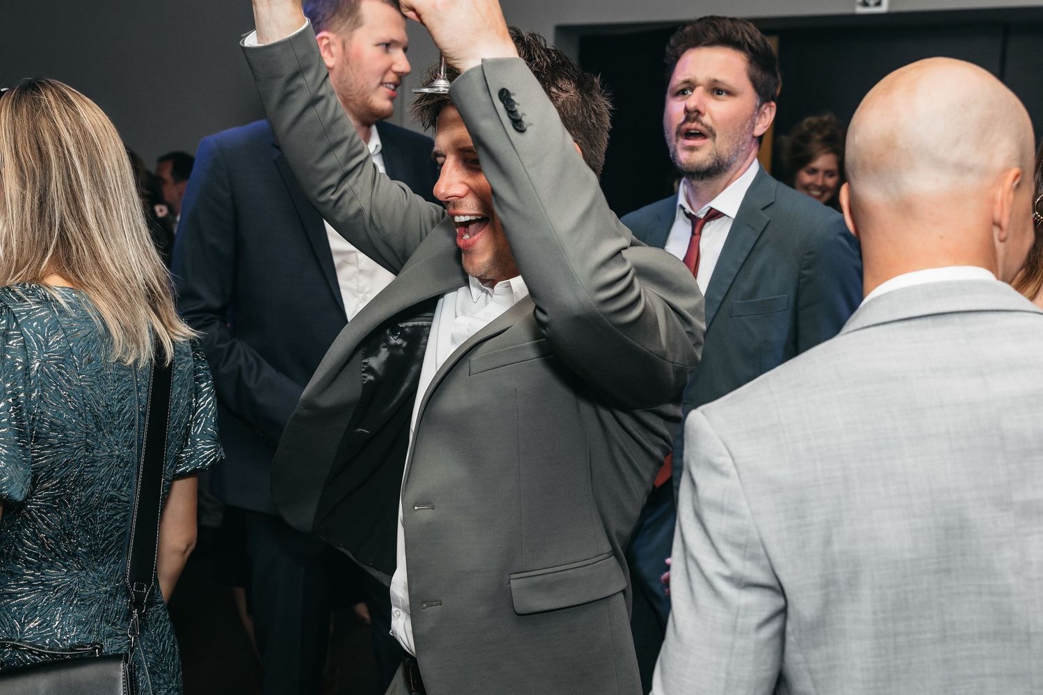 Excited man in grey suit dances with arms raised at wedding reception party.