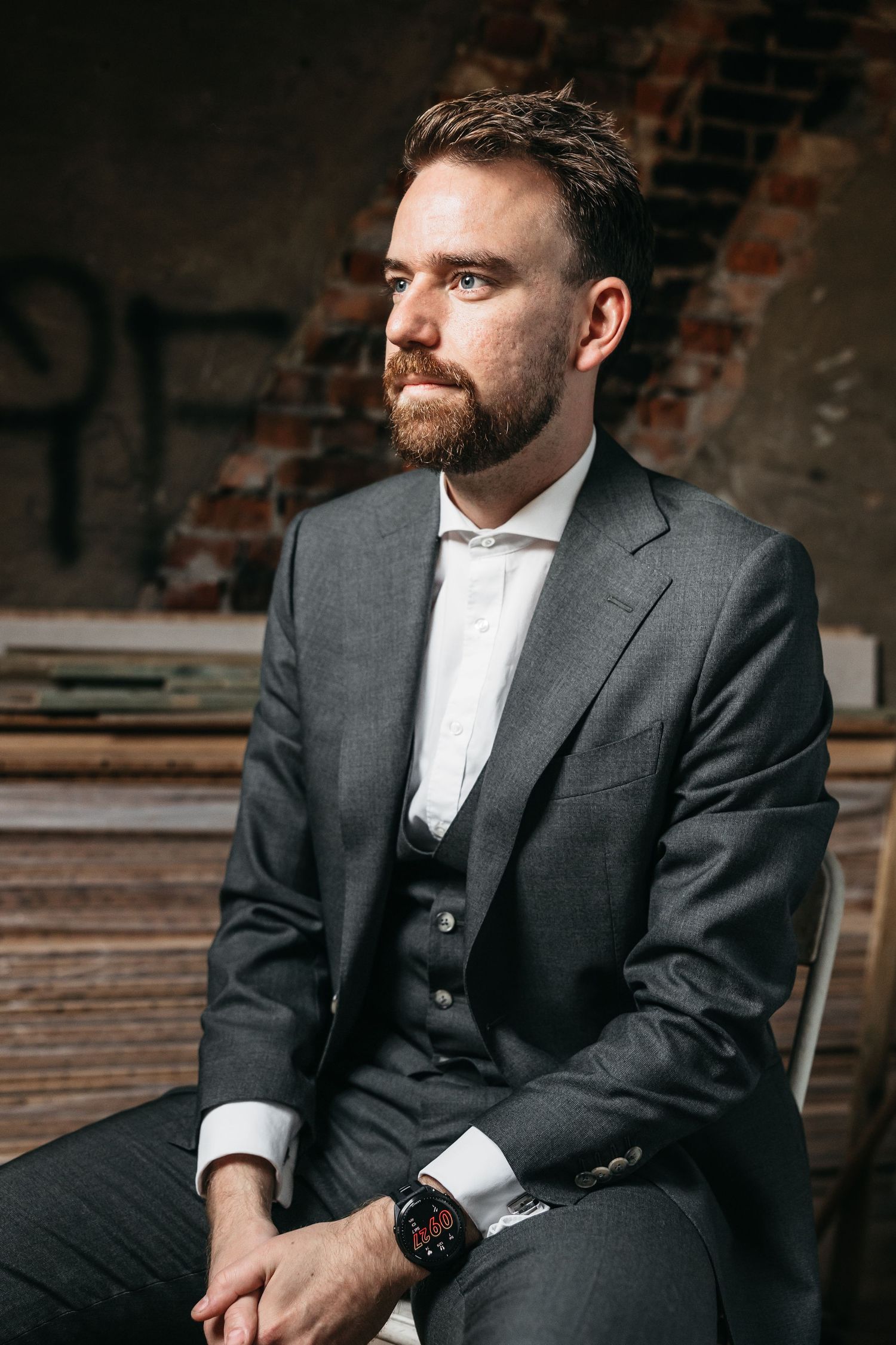 Bearded groom in gray three-piece suit with watch sitting against rustic brick wall backdrop.