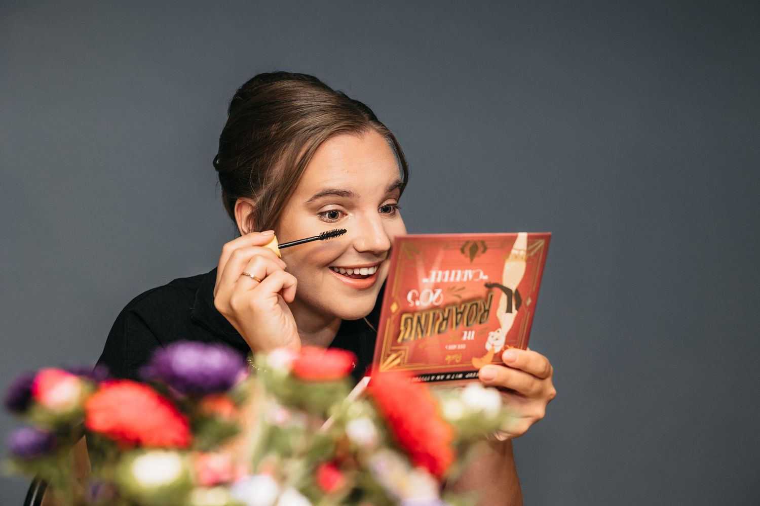Smiling young woman applying mascara while holding a makeup palette against a gray background.