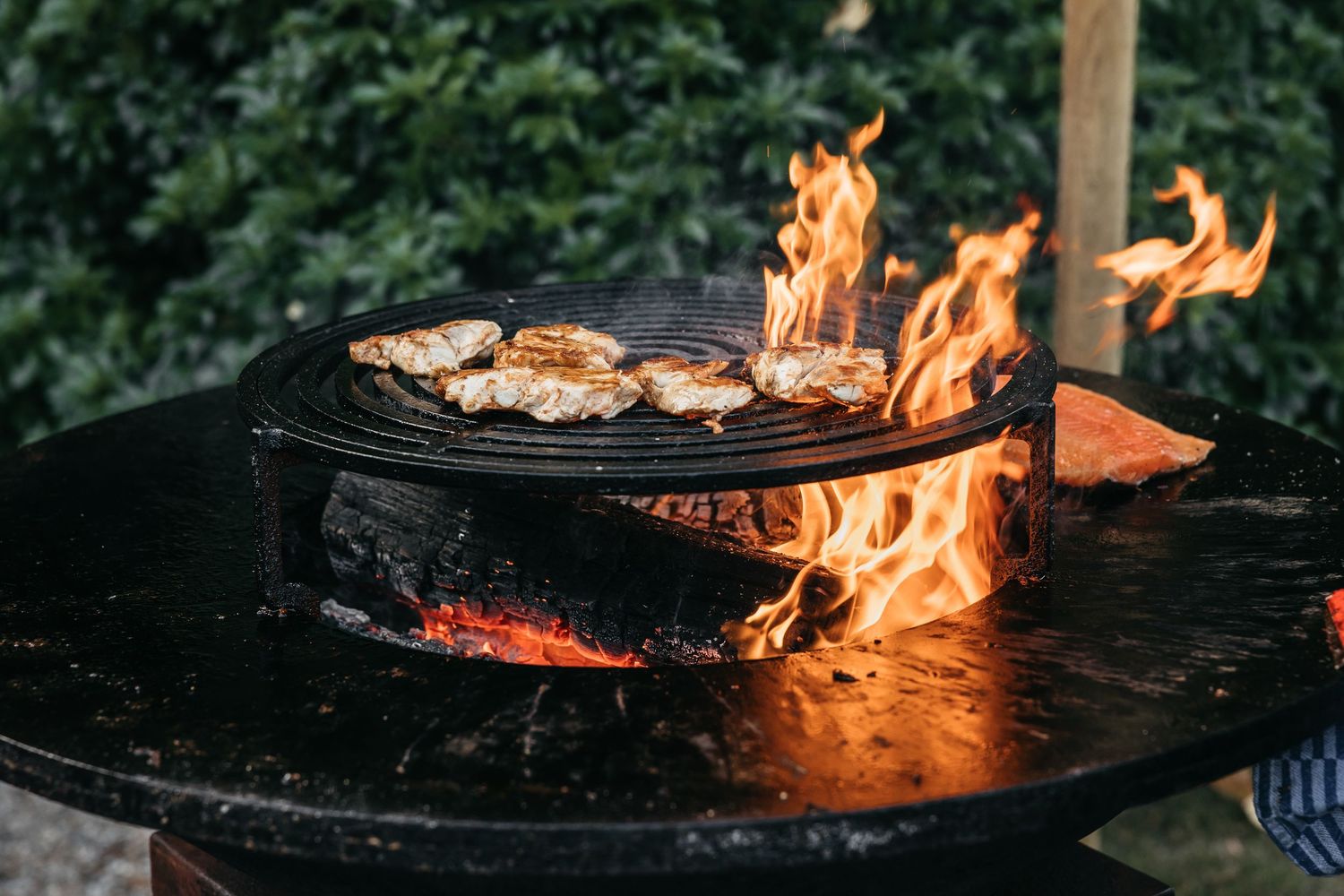 Meat grilling over open flames on large round outdoor fire pit BBQ grill at wedding reception.