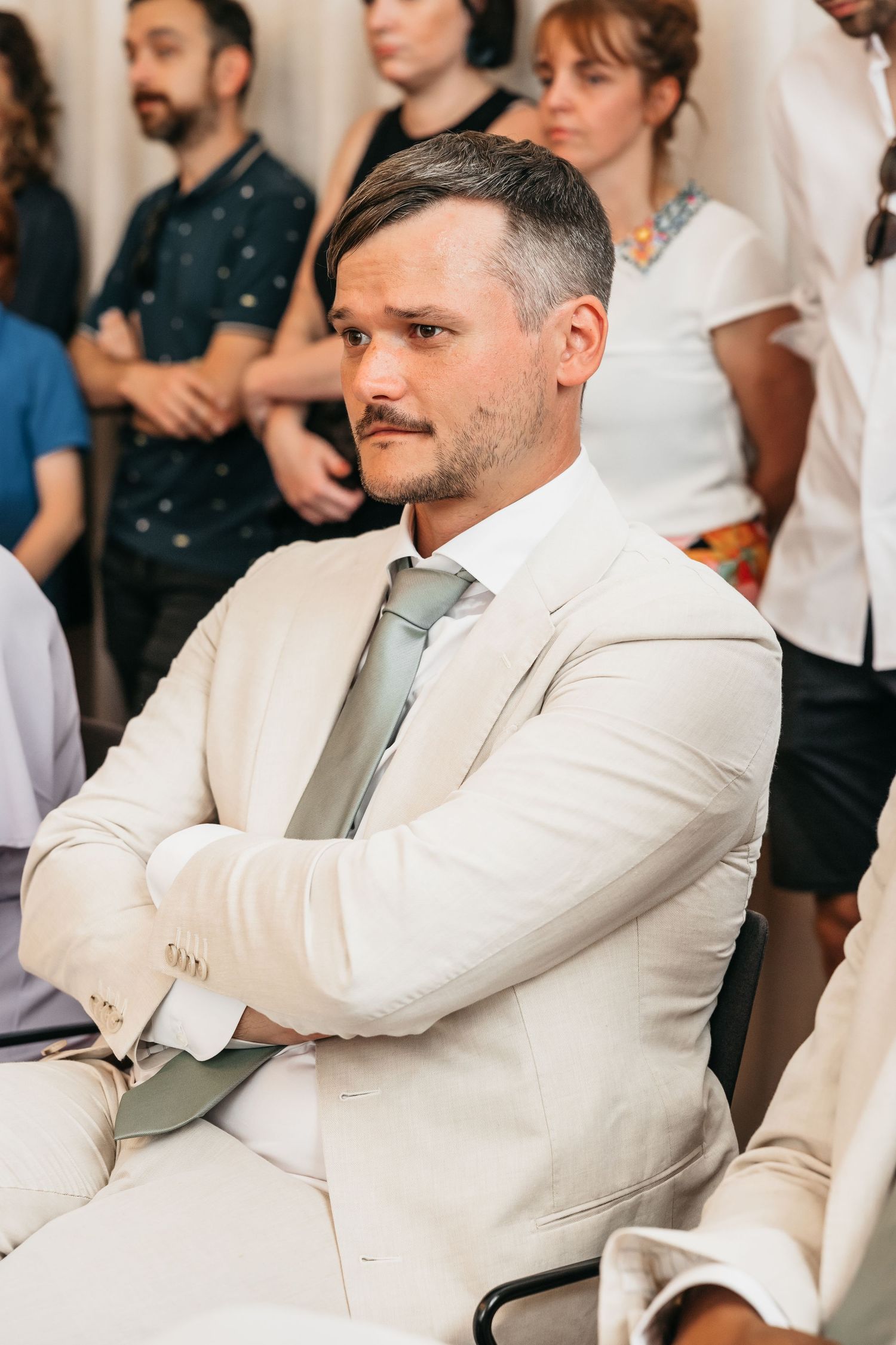 Elegantly dressed man in cream suit with arms crossed seated at wedding ceremony.