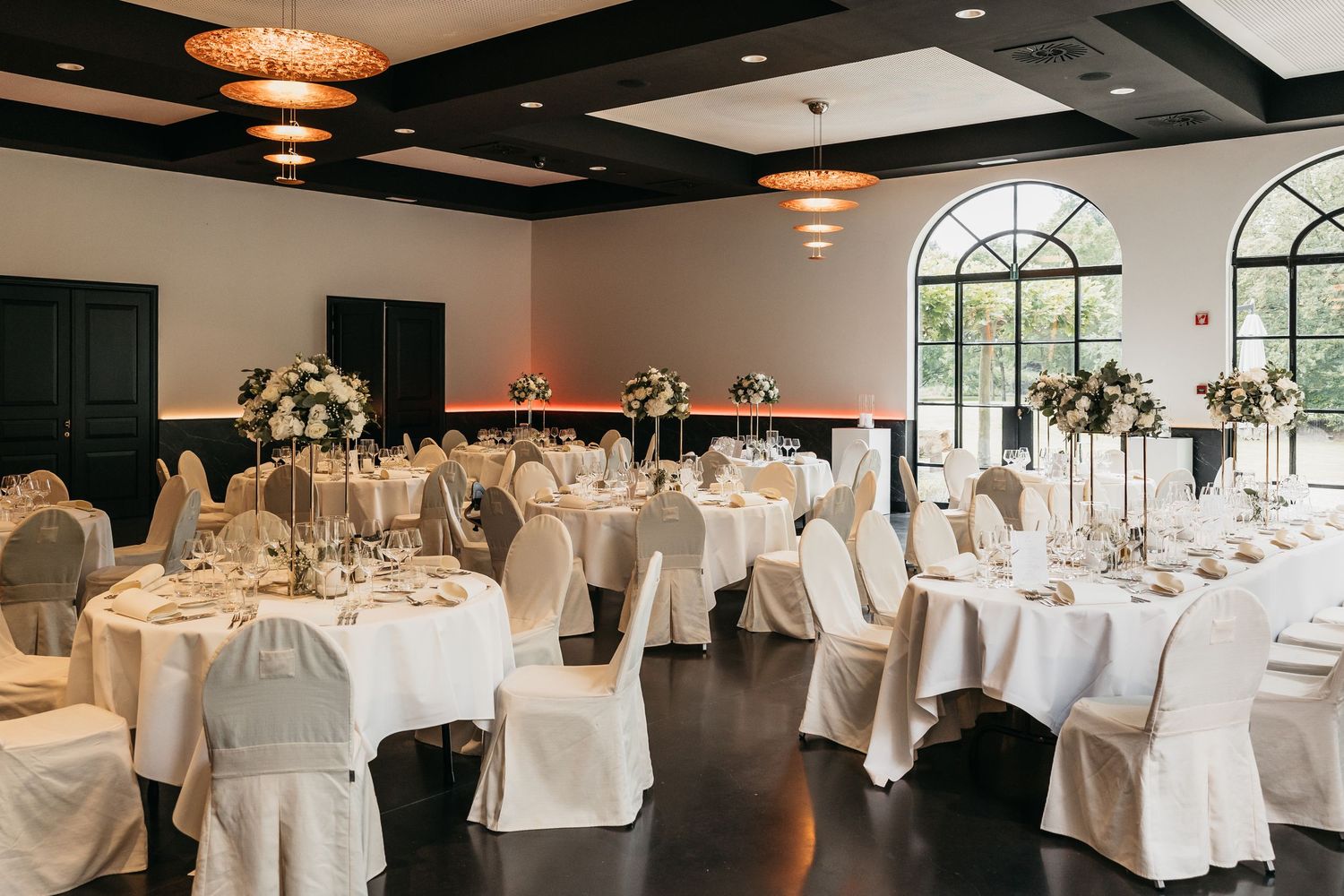 Sophisticated wedding banquet hall with white draped chairs, tall floral arrangements, and arched windows.