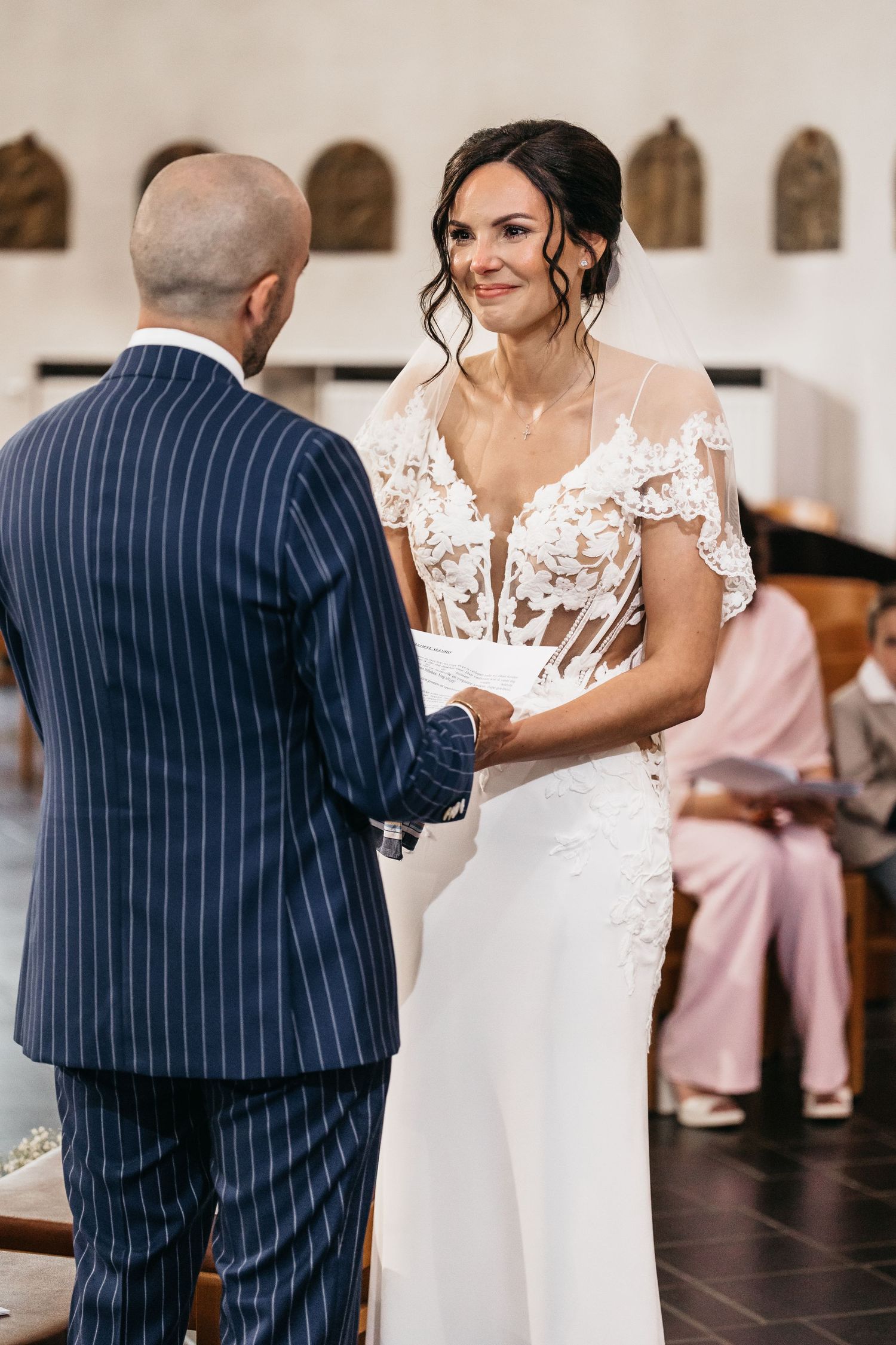 Smiling bride in lace wedding dress holds hands with groom at church altar.