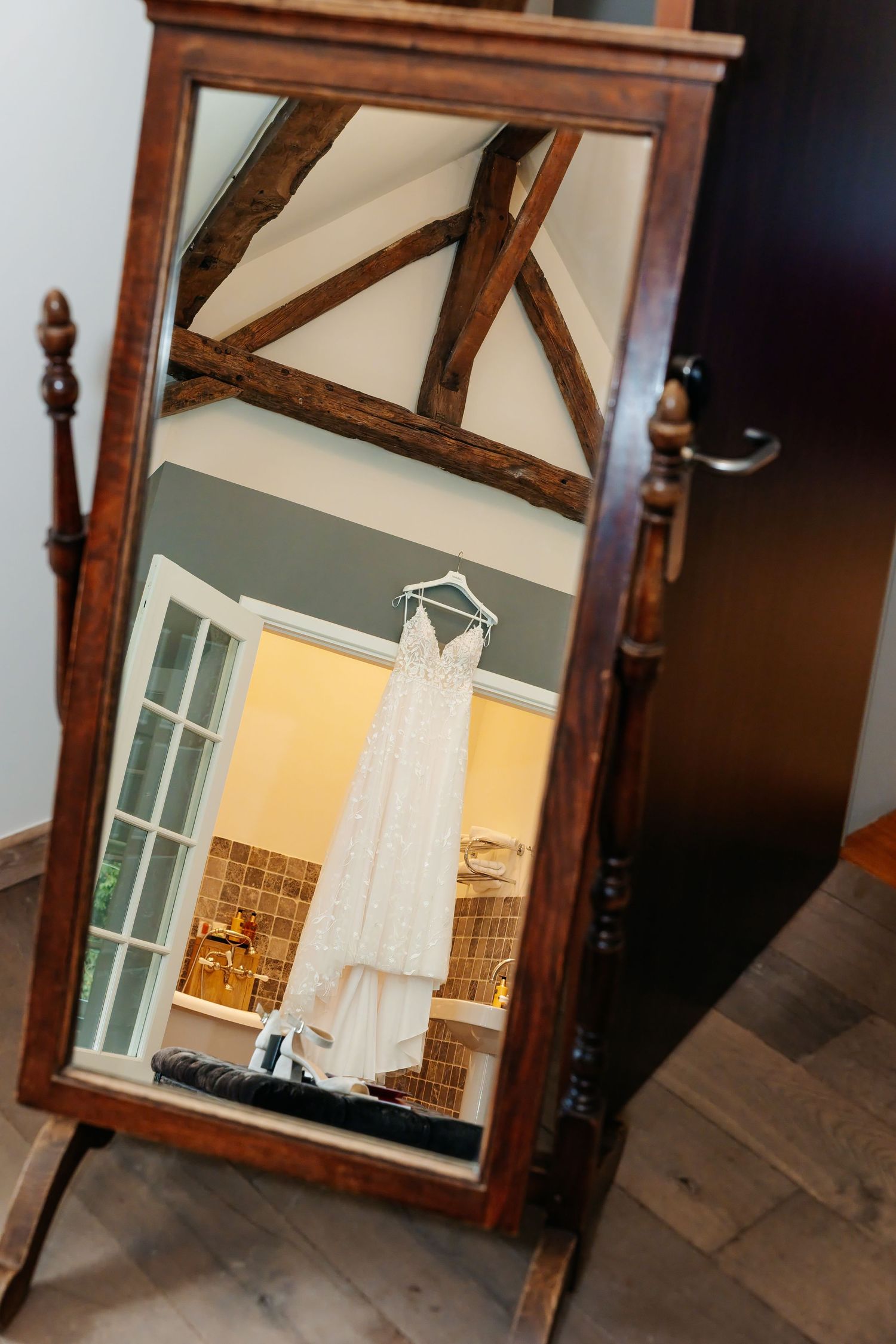 Elegant wooden cheval mirror reflecting a white lace wedding dress hanging in a rustic venue with exposed wooden beams.