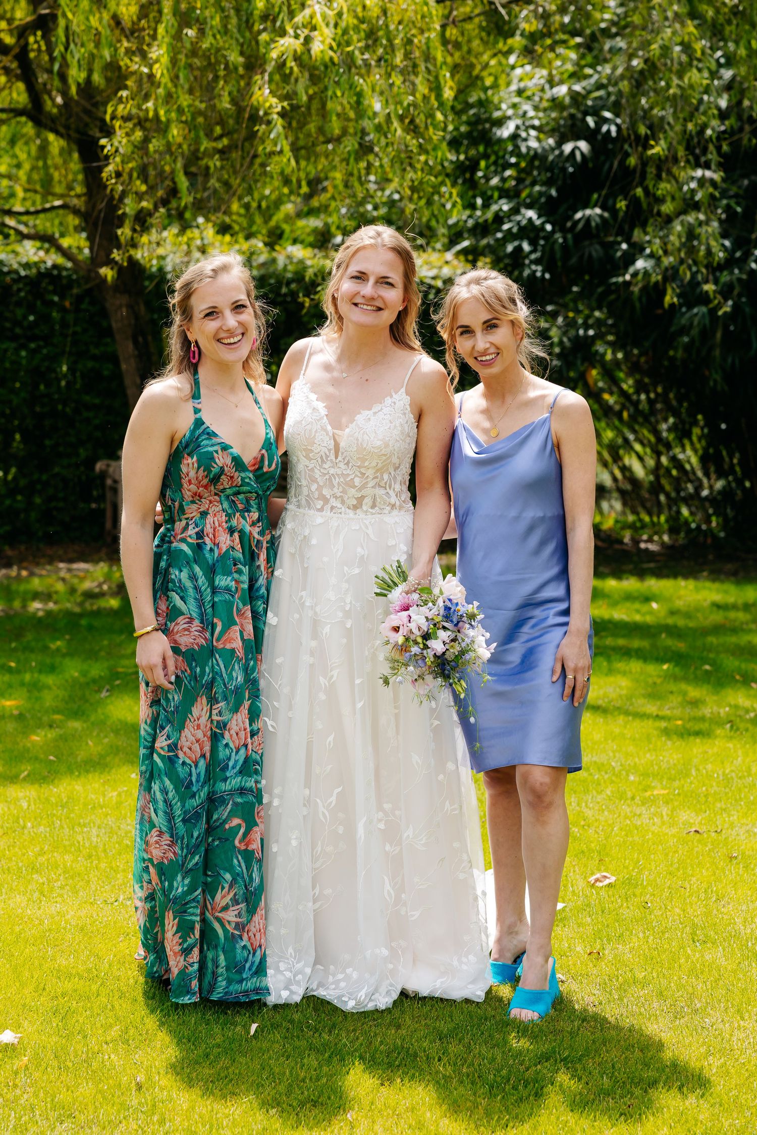 Bride in white lace gown poses with two guests in colorful dresses on green lawn.