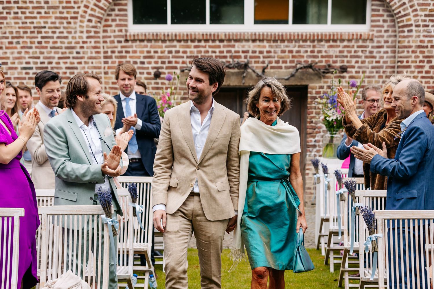 Groom walking with elegantly dressed woman through applauding wedding guests at outdoor ceremony.
