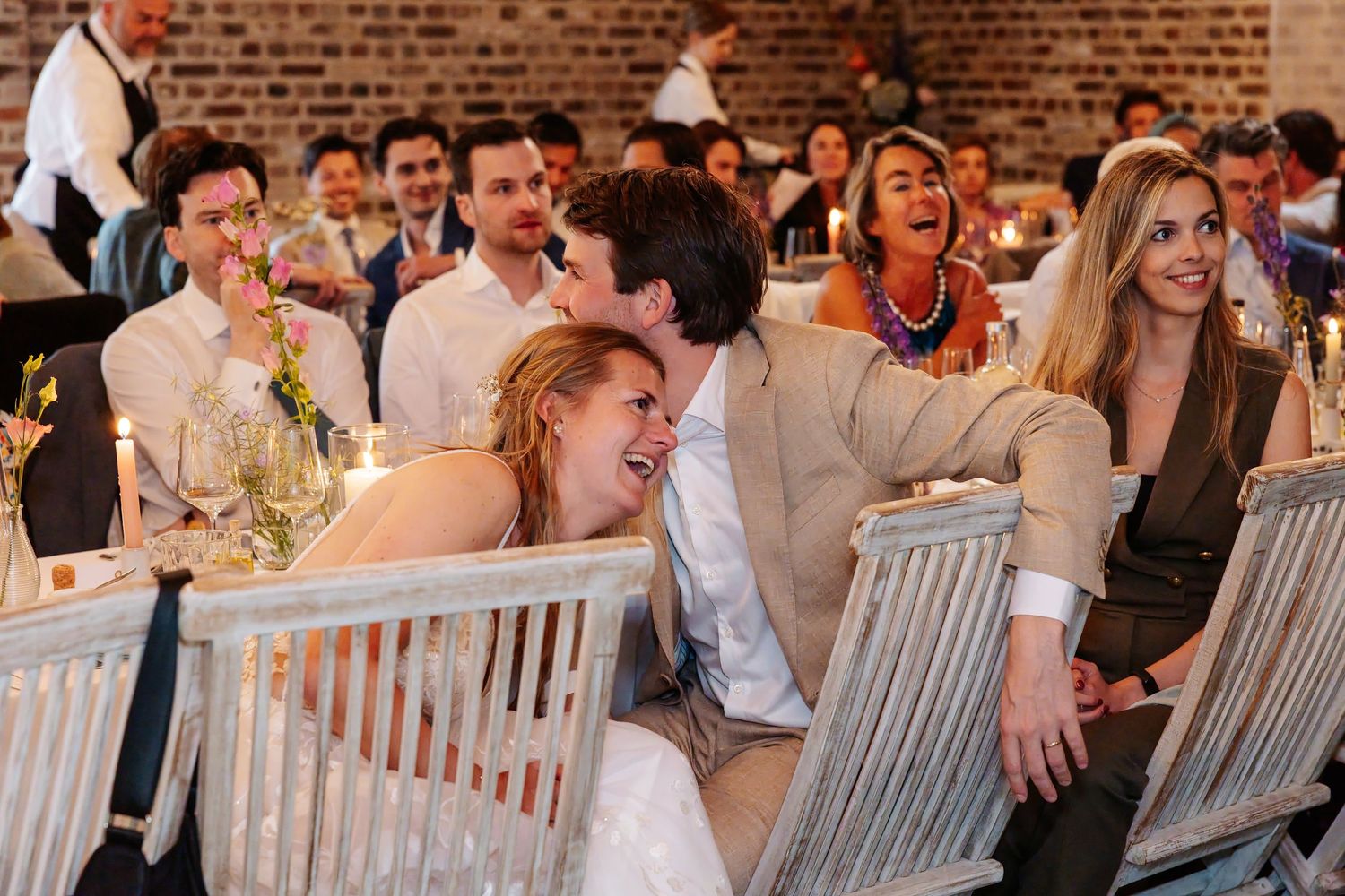 Bride and groom laugh joyfully with guests during lively wedding reception dinner at rustic venue.