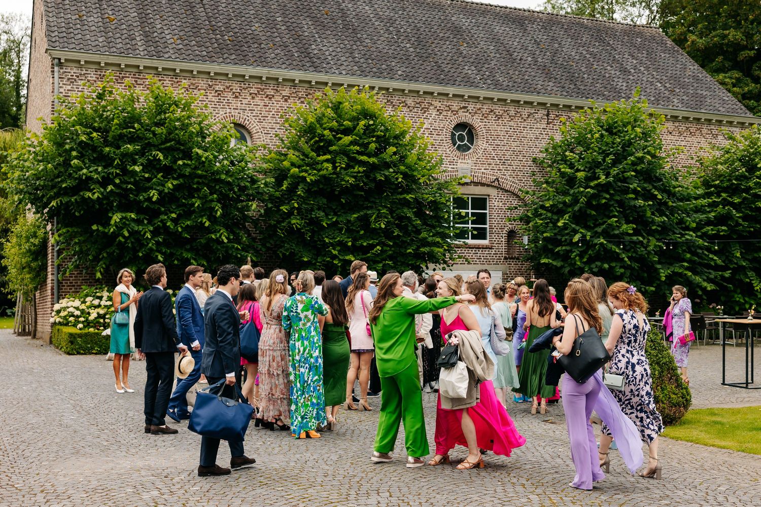 Wedding guests mingling outdoors in colorful attire on a cobblestone courtyard beside a historic brick building.