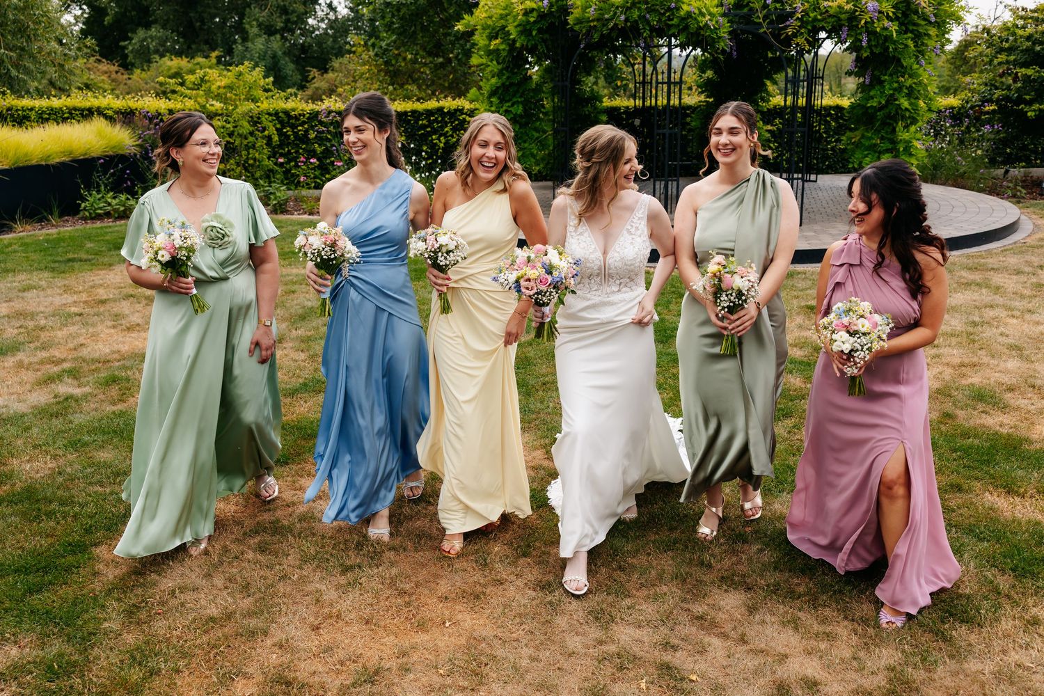 Bride in ivory gown walks laughing with five bridesmaids in colorful pastel mismatched dresses outdoors.