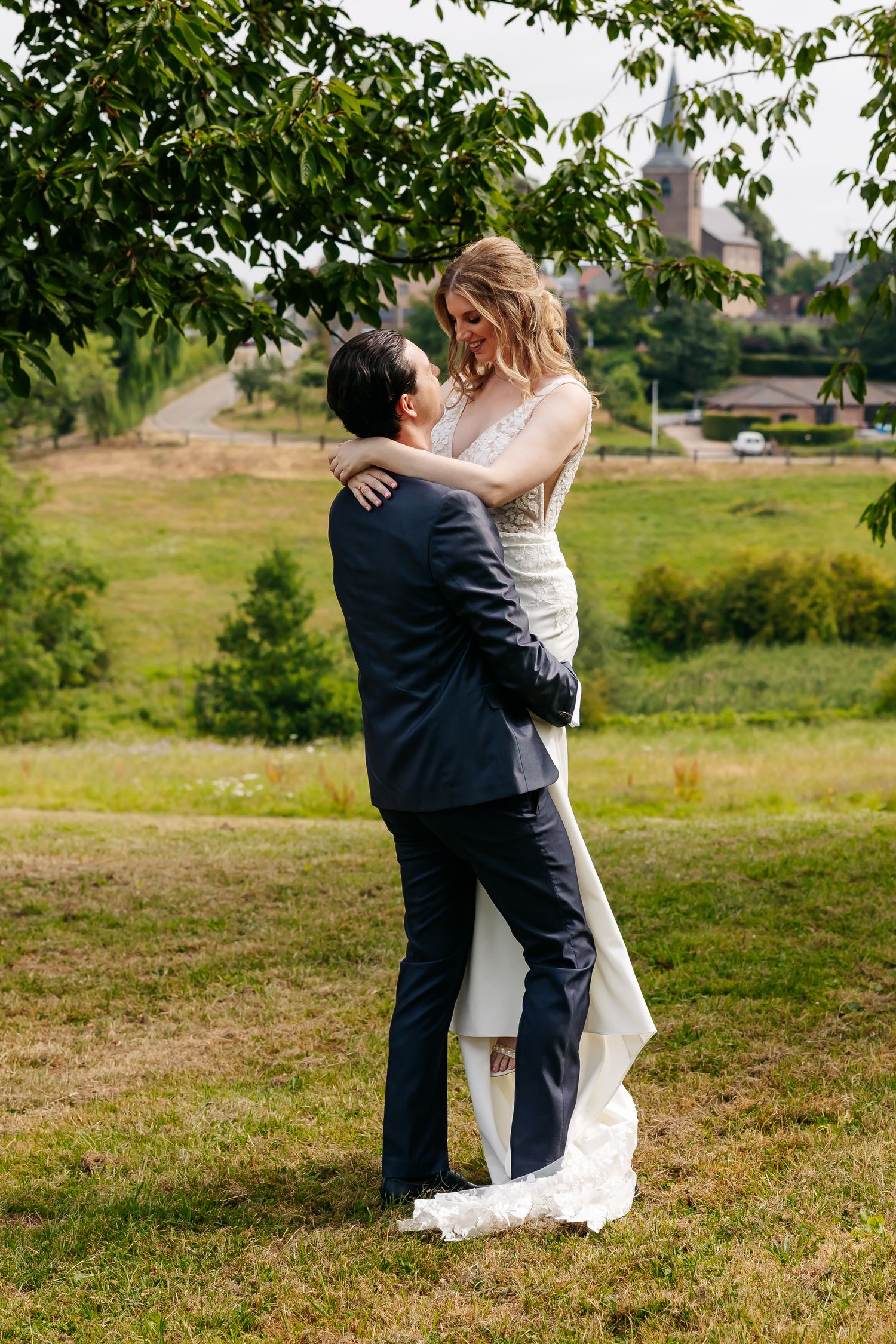 Groom lifting bride in white lace wedding dress while gazing at each other in green countryside.