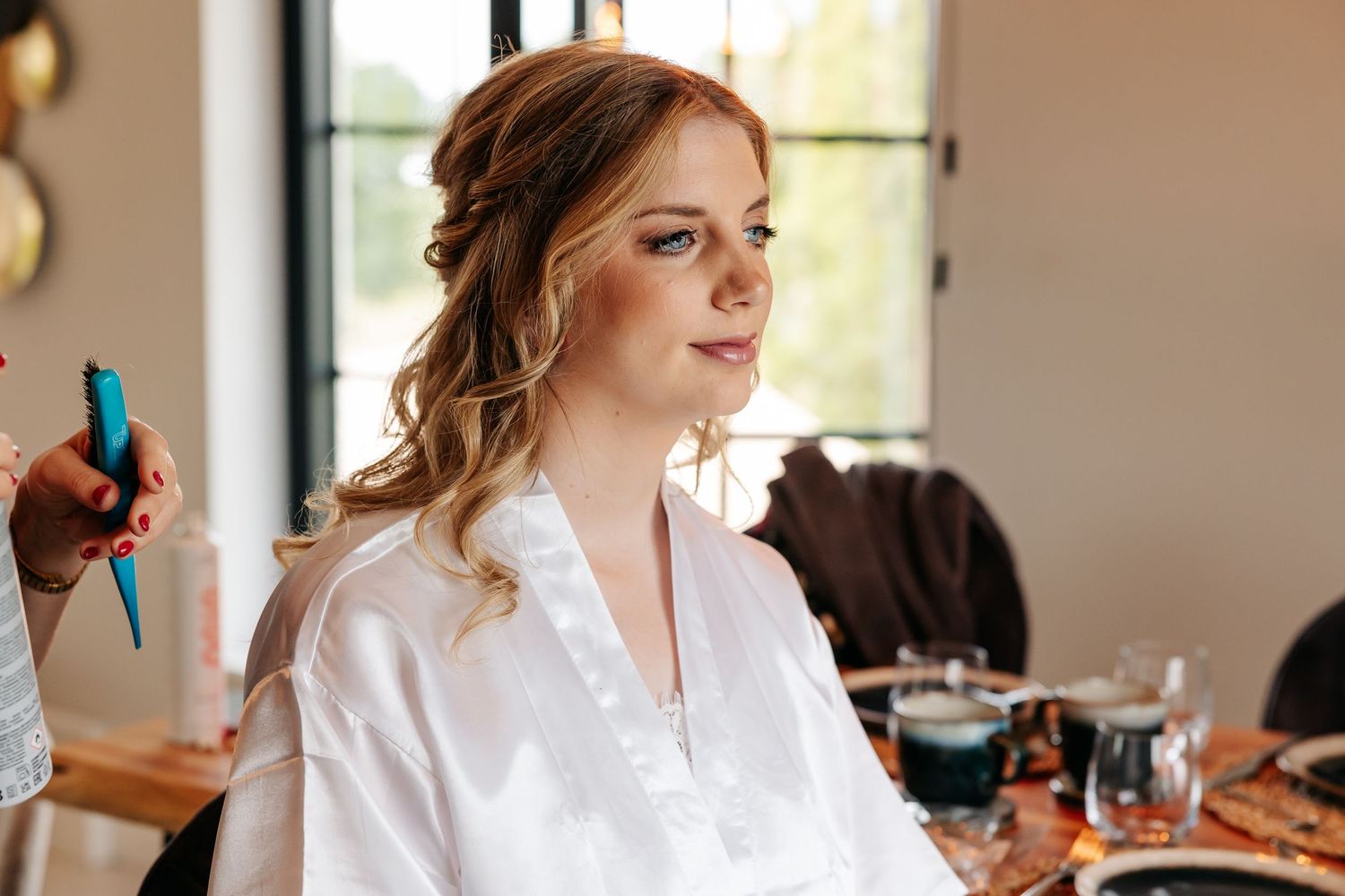 Bride in white robe sits patiently during hair and makeup application for her wedding day.