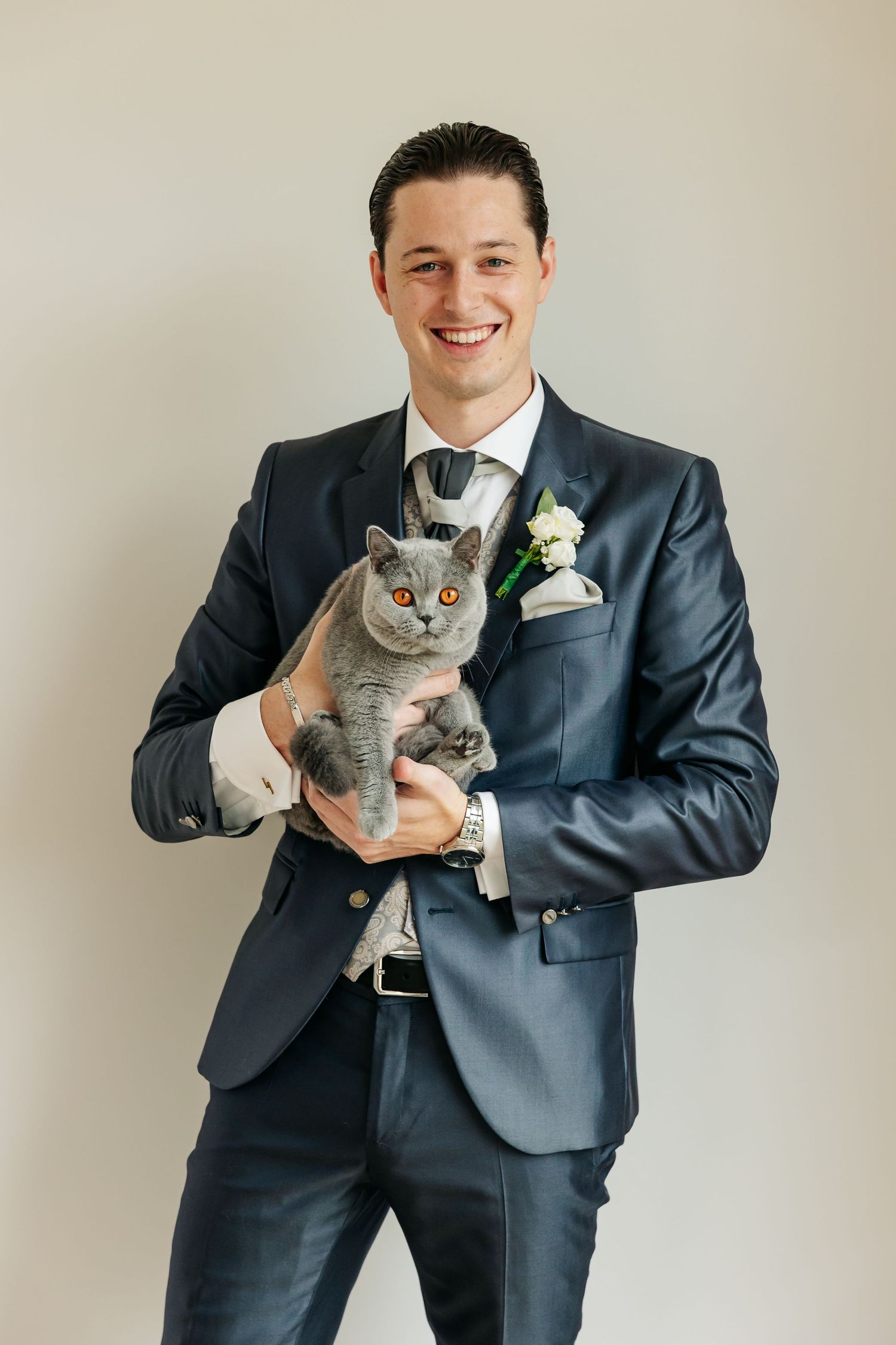 Smiling groom in navy suit holds gray British Shorthair cat with orange eyes on wedding day.