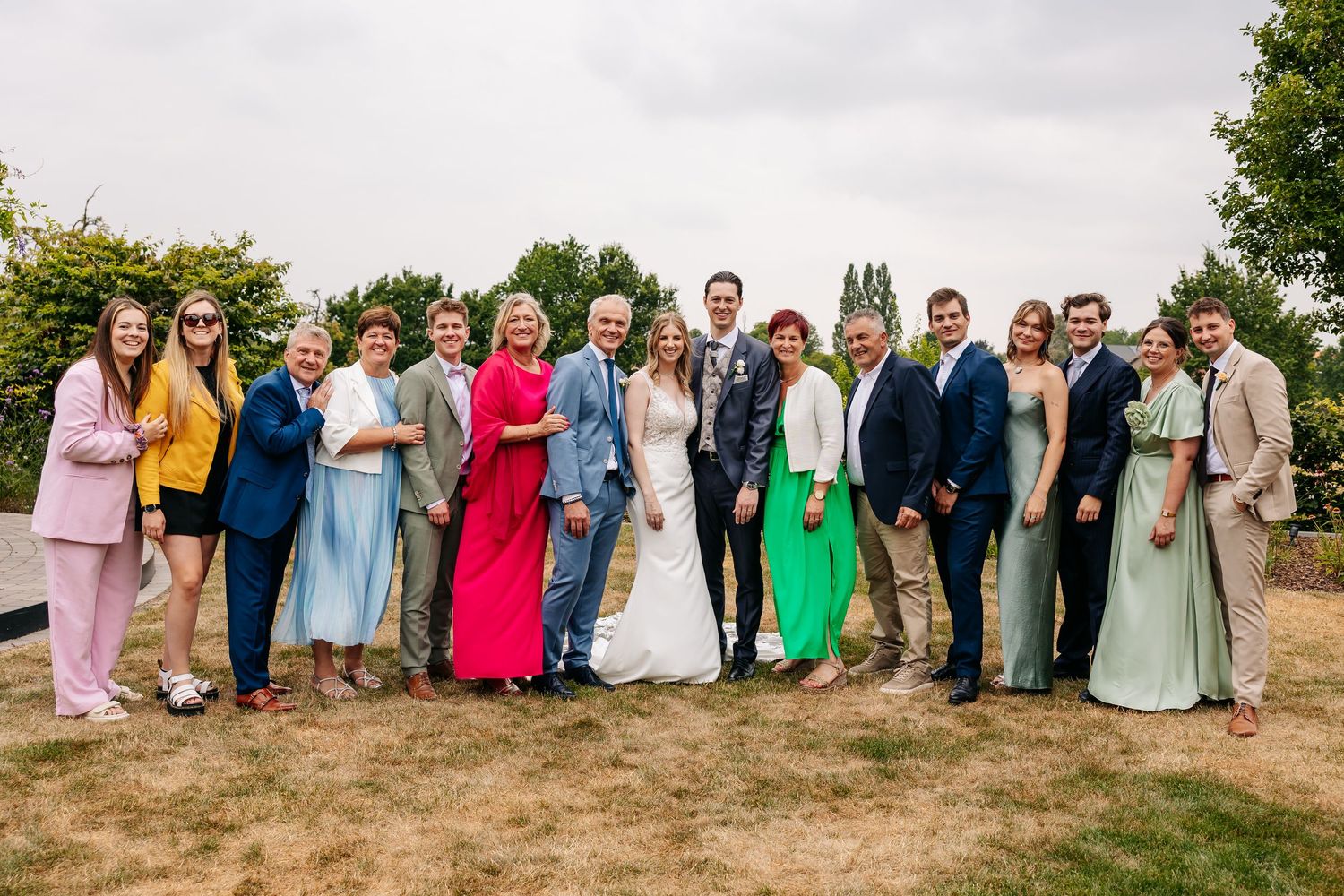 A large wedding party group portrait with bride and groom surrounded by family and friends outdoors.