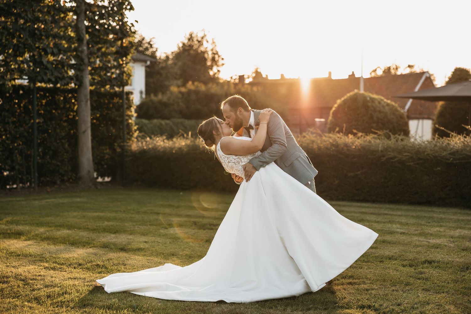 Groom dipping bride in white gown during romantic sunset wedding photo in garden.