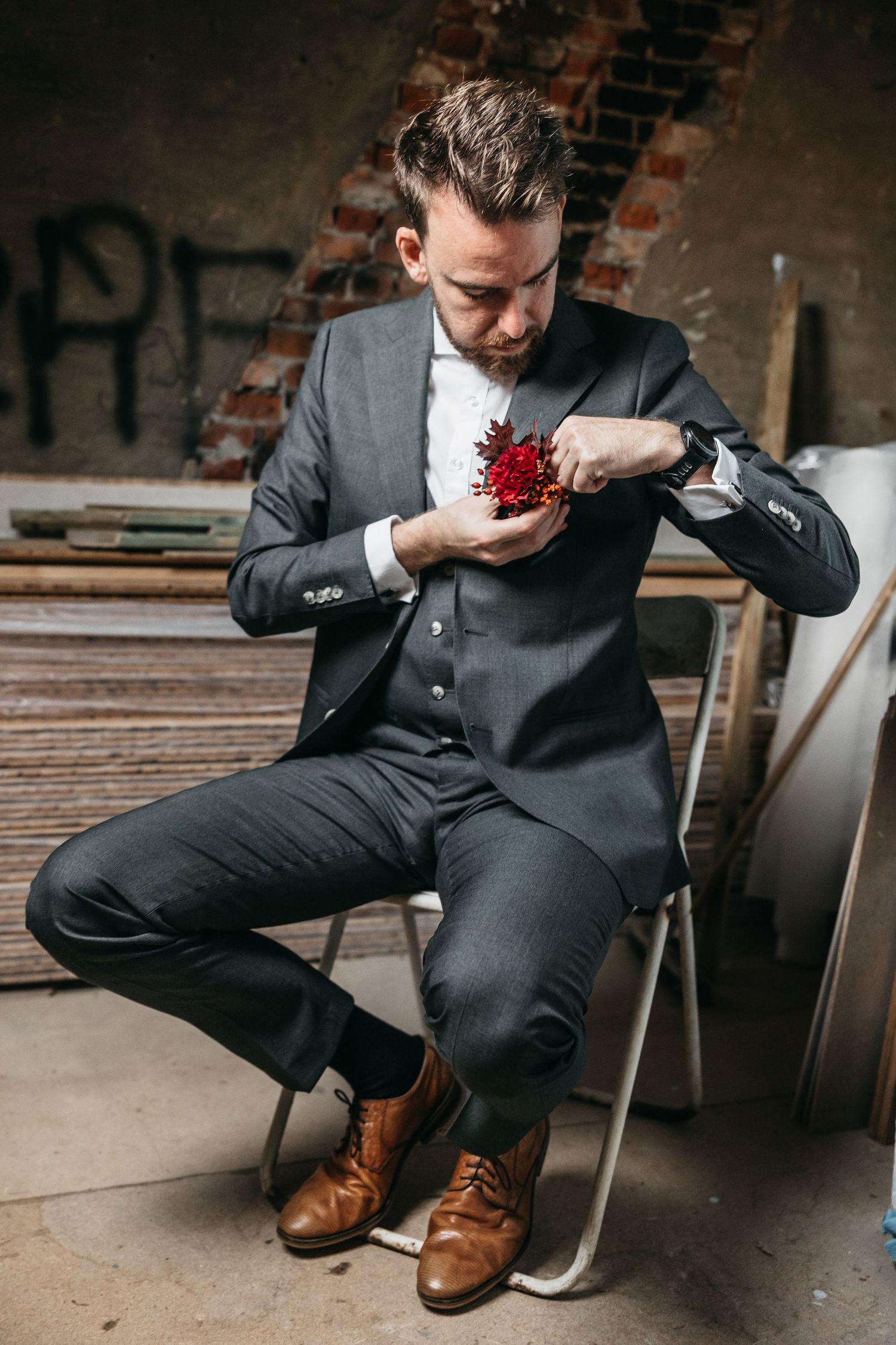 Groom in gray suit attaching red floral boutonniere to lapel in rustic warehouse venue.