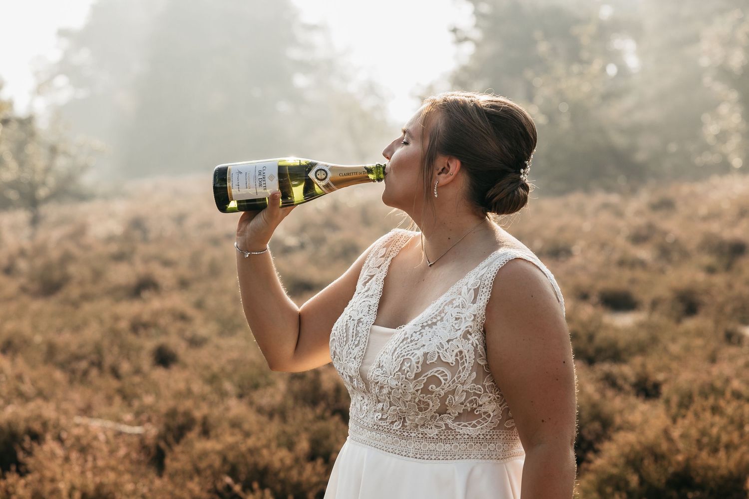 Bride in white lace wedding dress drinking champagne from bottle in sun-drenched heather field.