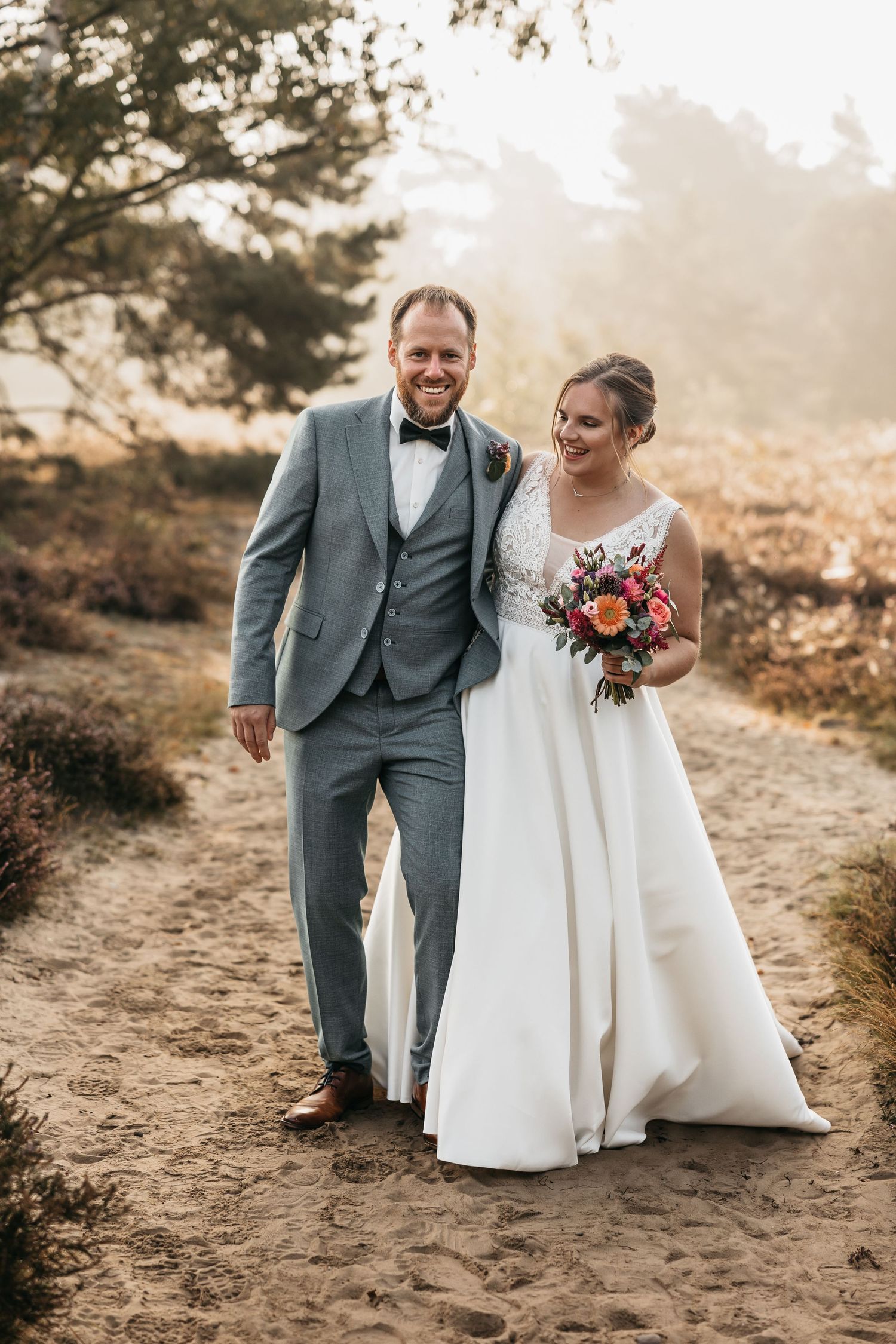 Smiling bride and groom walking hand in hand on a sandy path surrounded by heathland in soft golden light.