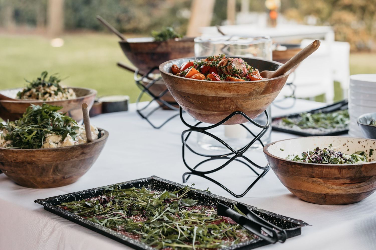 Elegant outdoor buffet spread with wooden bowls of fresh salads and garnished dishes on a white tablecloth.