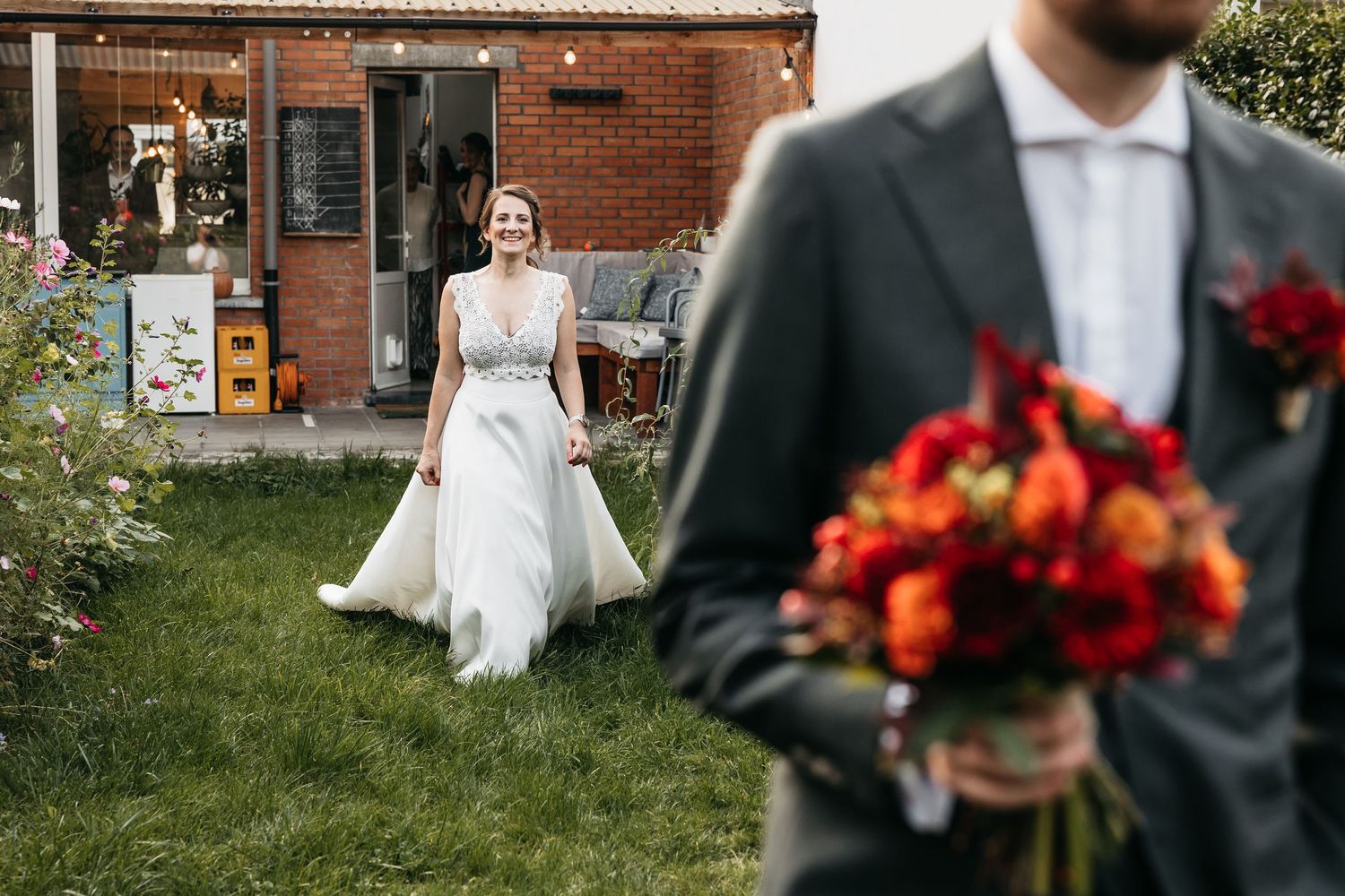Bride in white gown smiling and walking toward groom holding a red bouquet in a garden.