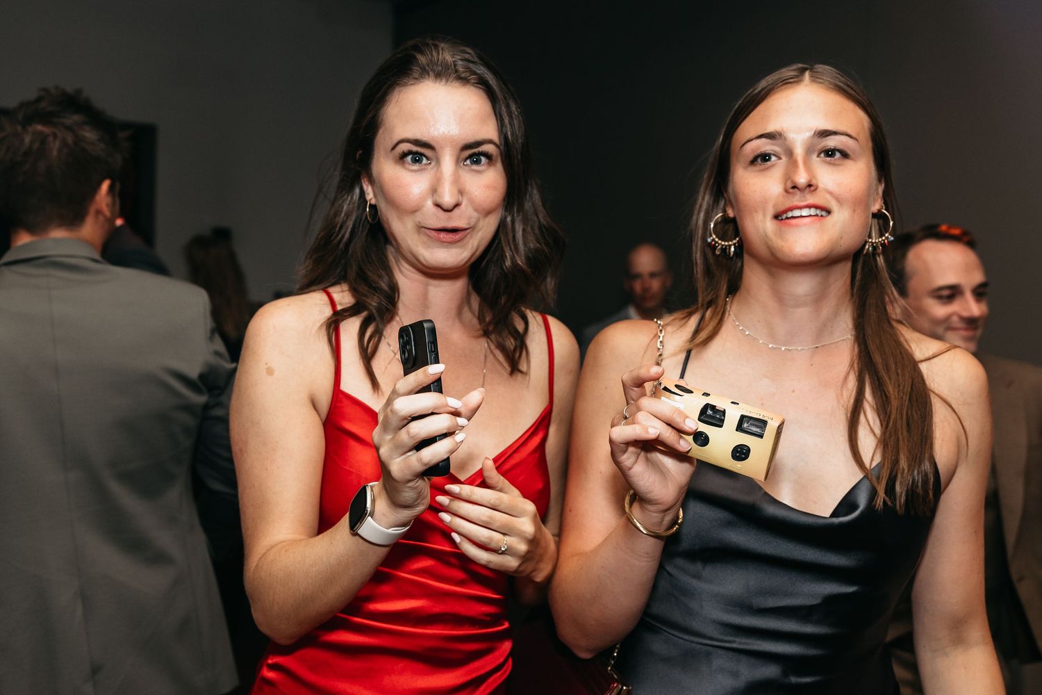 Two women in red and black dresses pose together holding cameras at wedding.
