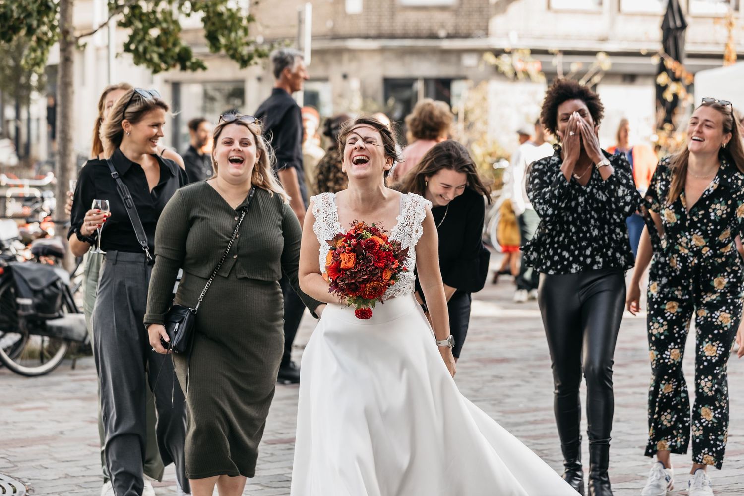 Laughing bride walking through city streets with bridesmaids holding a red bridal bouquet.
