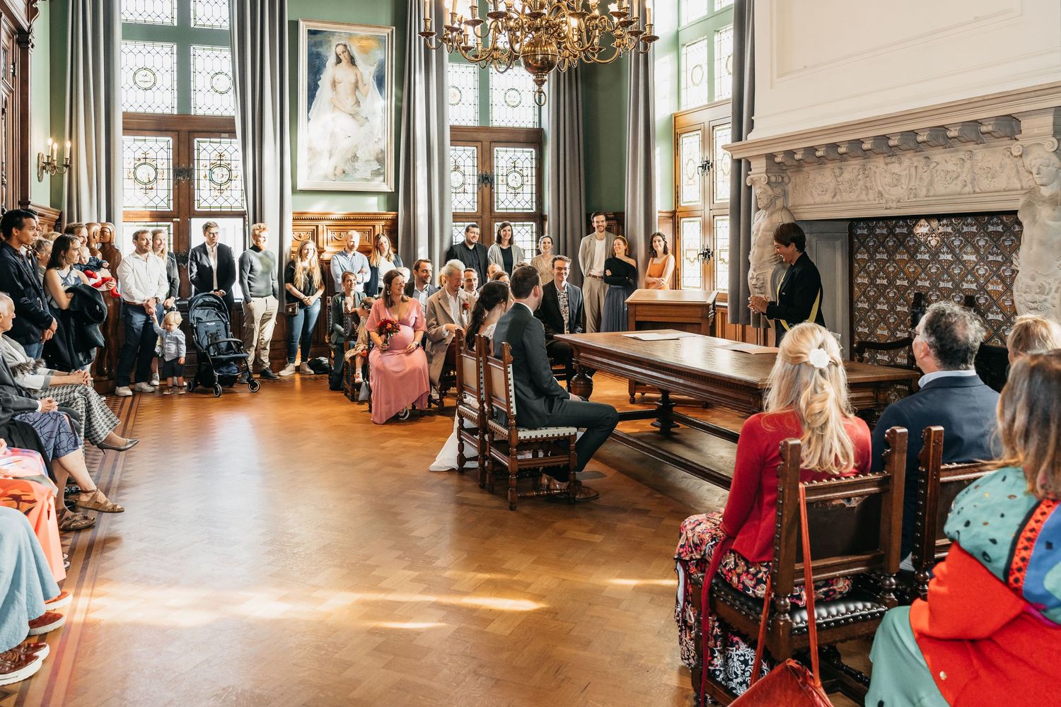 Wide view of a formal wedding ceremony in a grand historic room with stained glass windows and ornate chandelier.