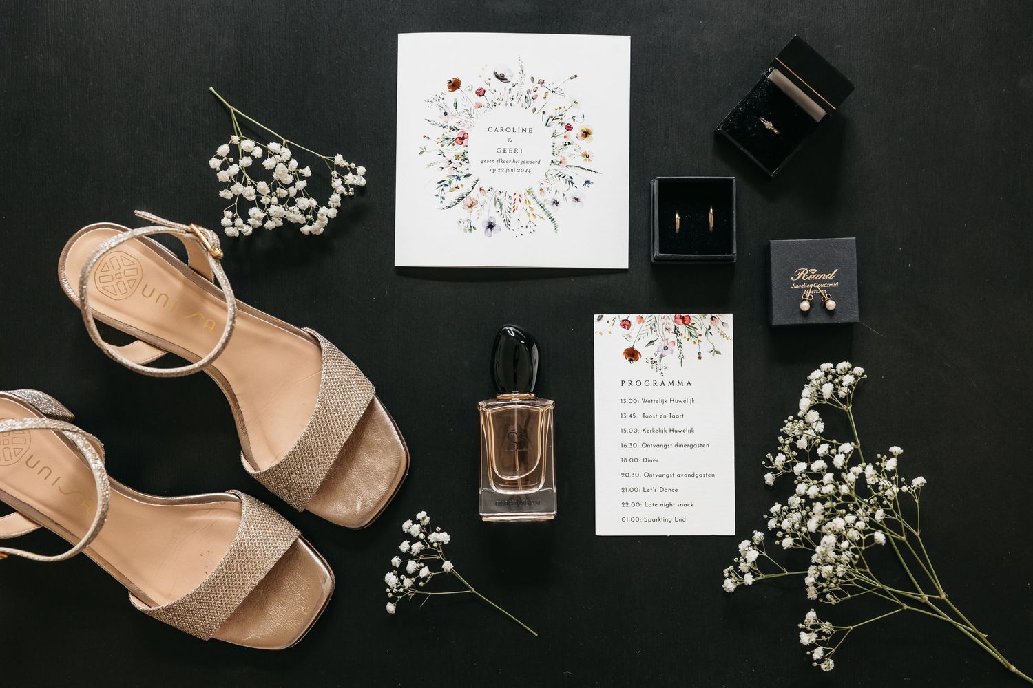 Flat lay of wedding accessories including gold heels, floral invitation, perfume, and baby's breath.