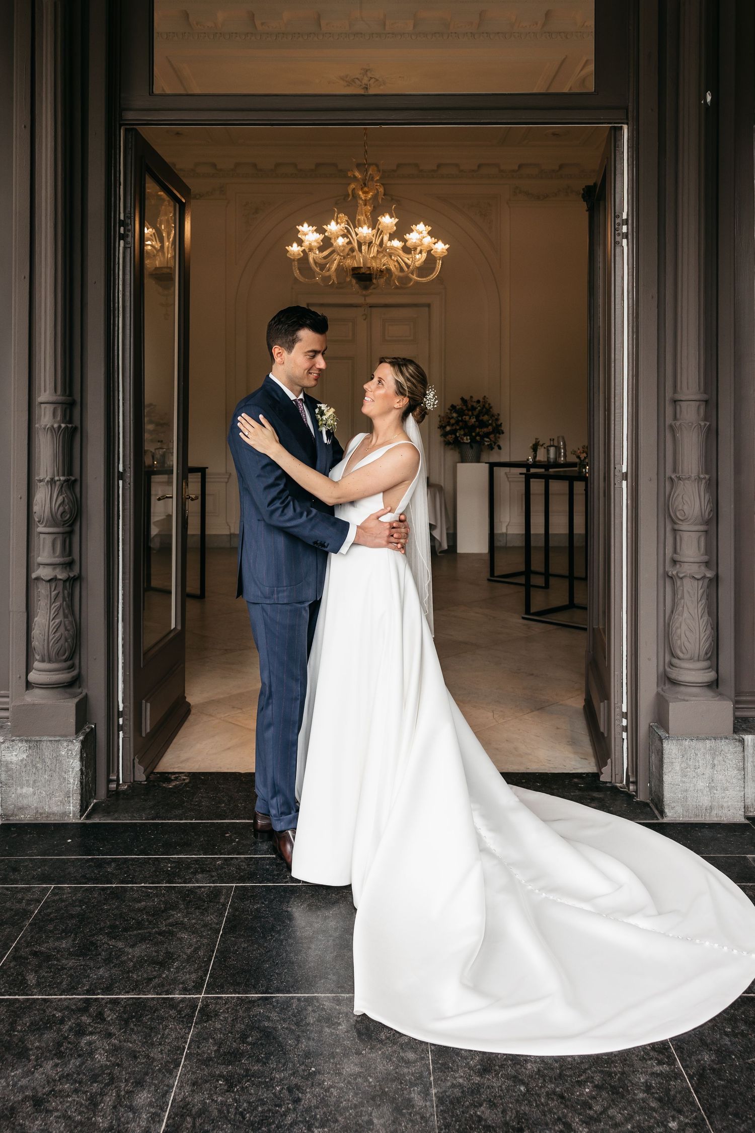 Bride and groom embrace romantically in grand doorway of elegant venue with chandelier glowing behind.
