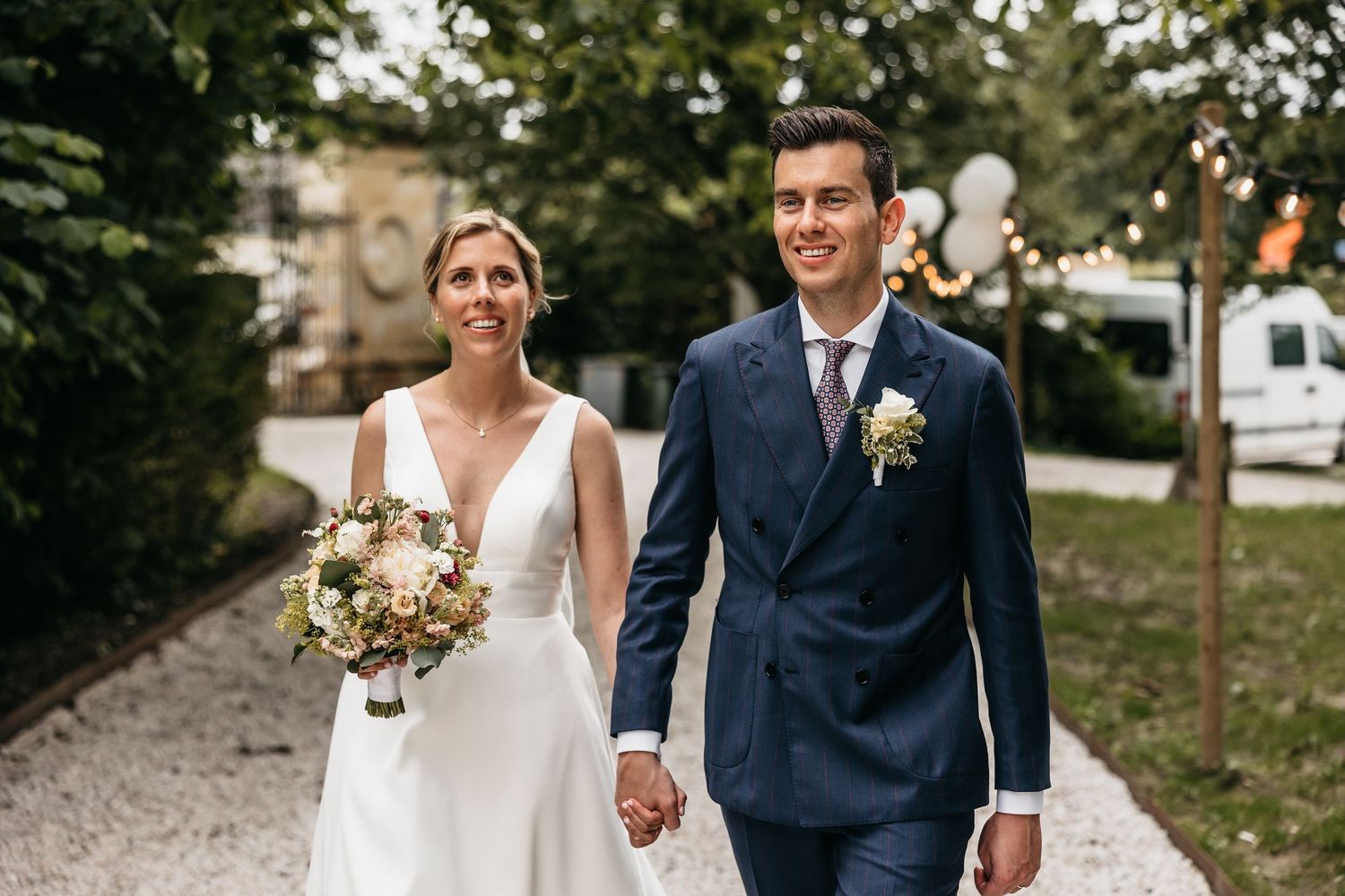 Happy bride and groom walking hand in hand along tree-lined path on their wedding day.