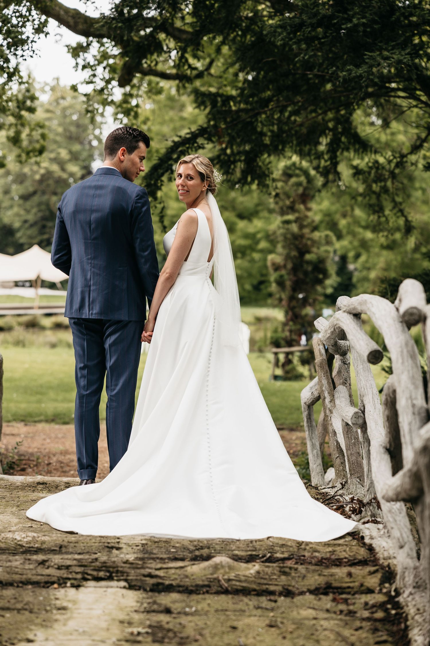 Bride in white gown and groom in navy suit holding hands on a rustic wooden bridge.