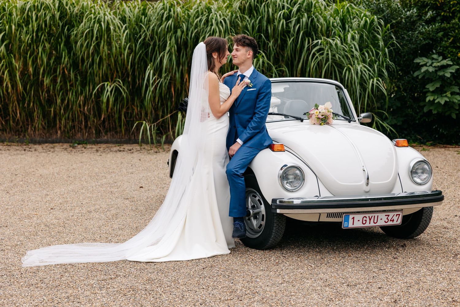 Bride and groom kissing beside a vintage white VW Beetle convertible on their wedding day.