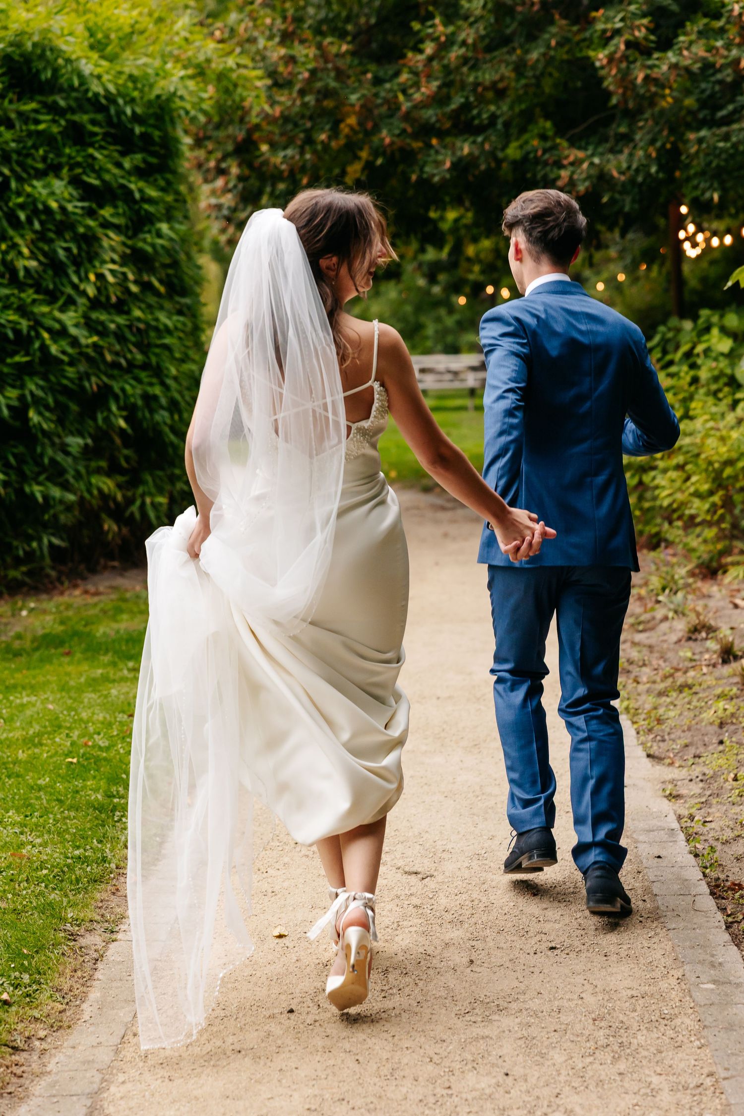 Back view of a bride holding her wedding dress and groom in blue suit walking hand in hand down a garden path.