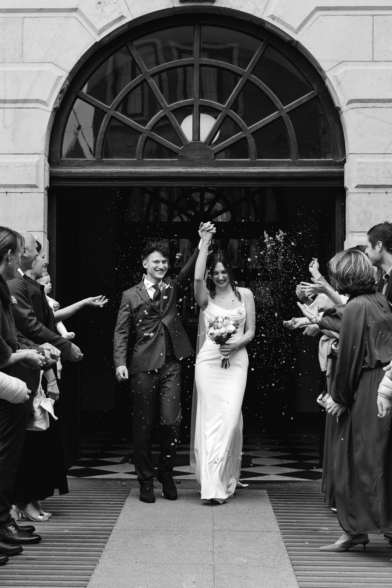 Bride and groom raise hands in triumph as confetti falls outside church in black and white.