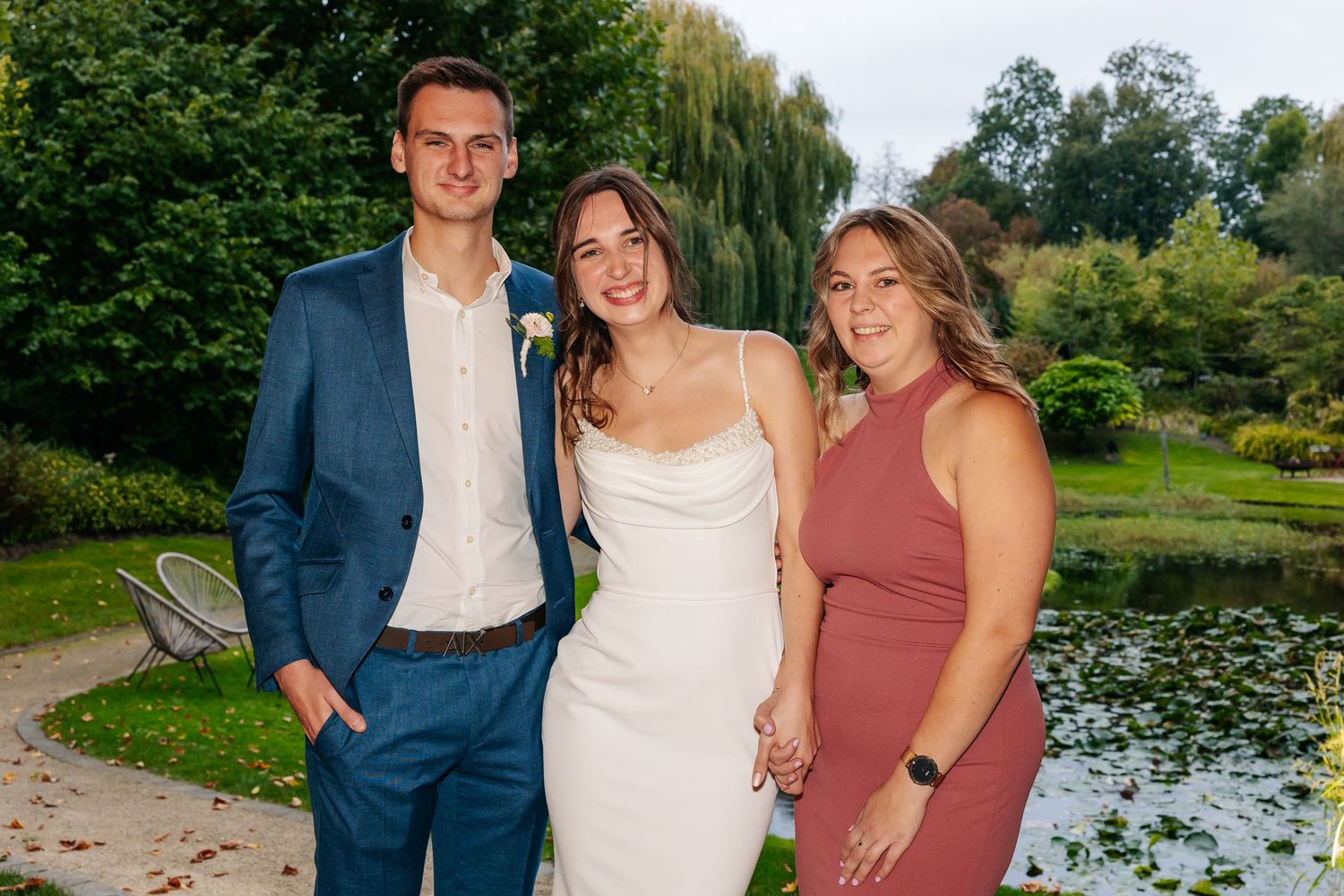 Bride in white gown posing with groom in blue suit and bridesmaid by garden pond.