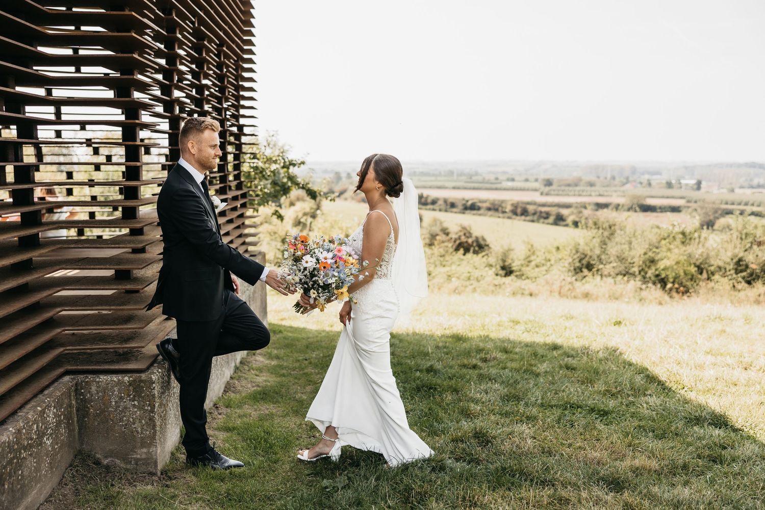 Bride holding a colorful bouquet walks with groom beside a rustic wooden structure overlooking green fields.