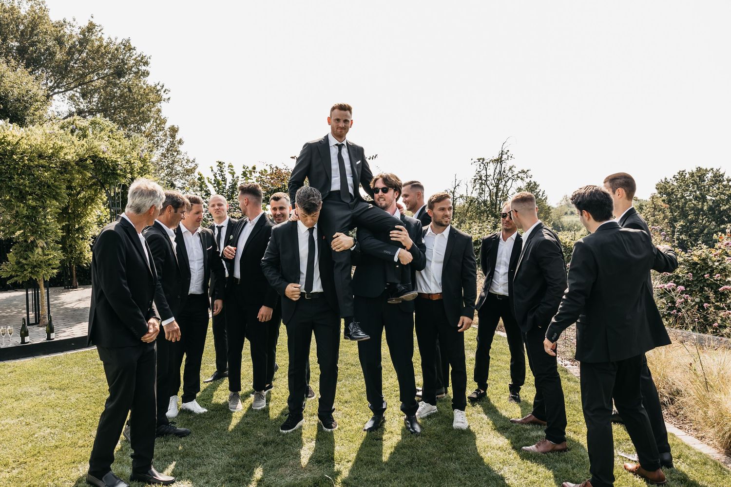 Groomsmen lifting groom on shoulders while group celebrates outdoors at sunny wedding.